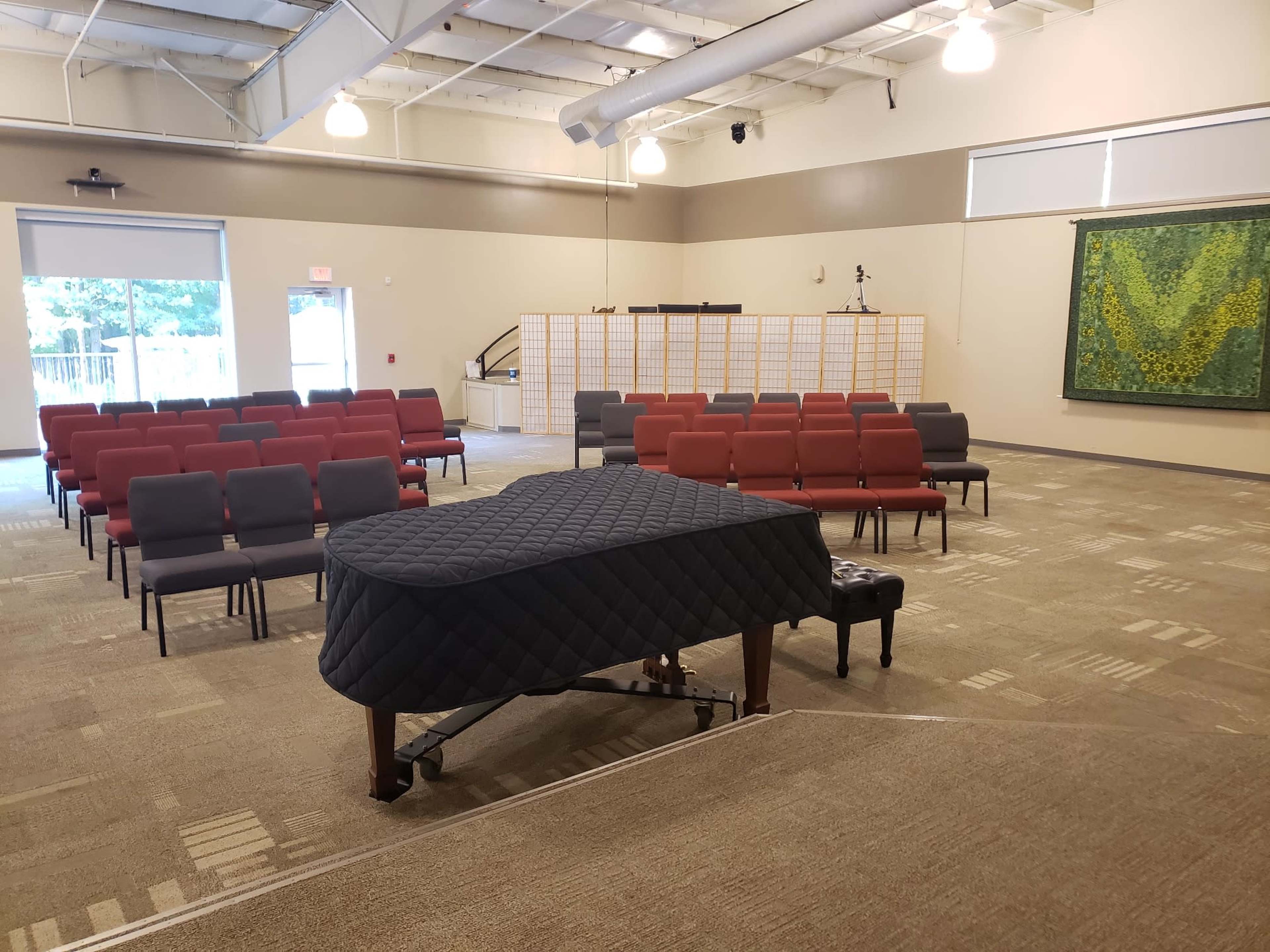 A grand piano covered with a protective cloth is positioned in front of a set of empty chairs arranged in rows in a spacious room.