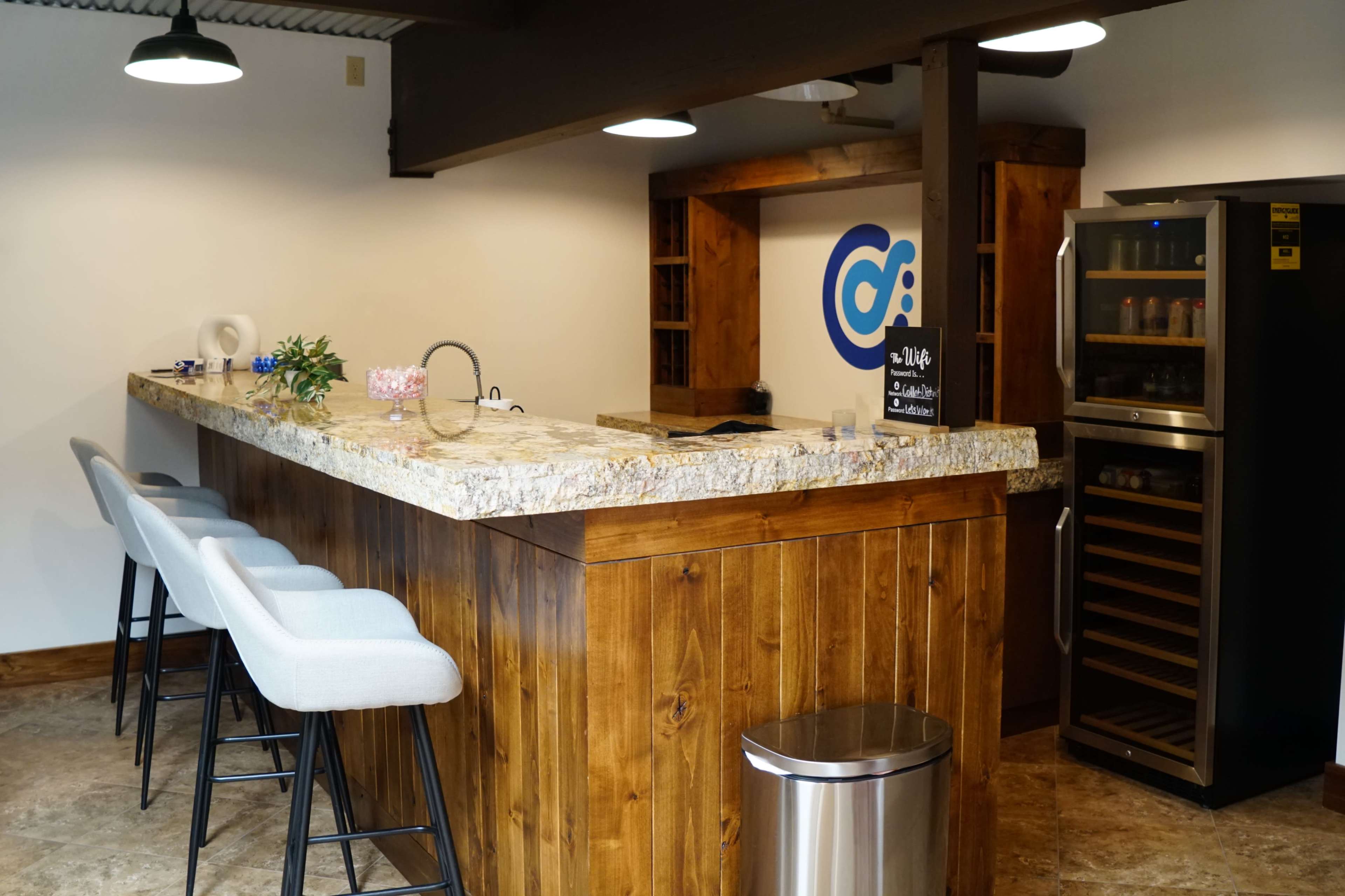 A minimalist bar area with a granite countertop, wooden cabinetry, a modern refrigerator, and several bar stools arranged along the counter.