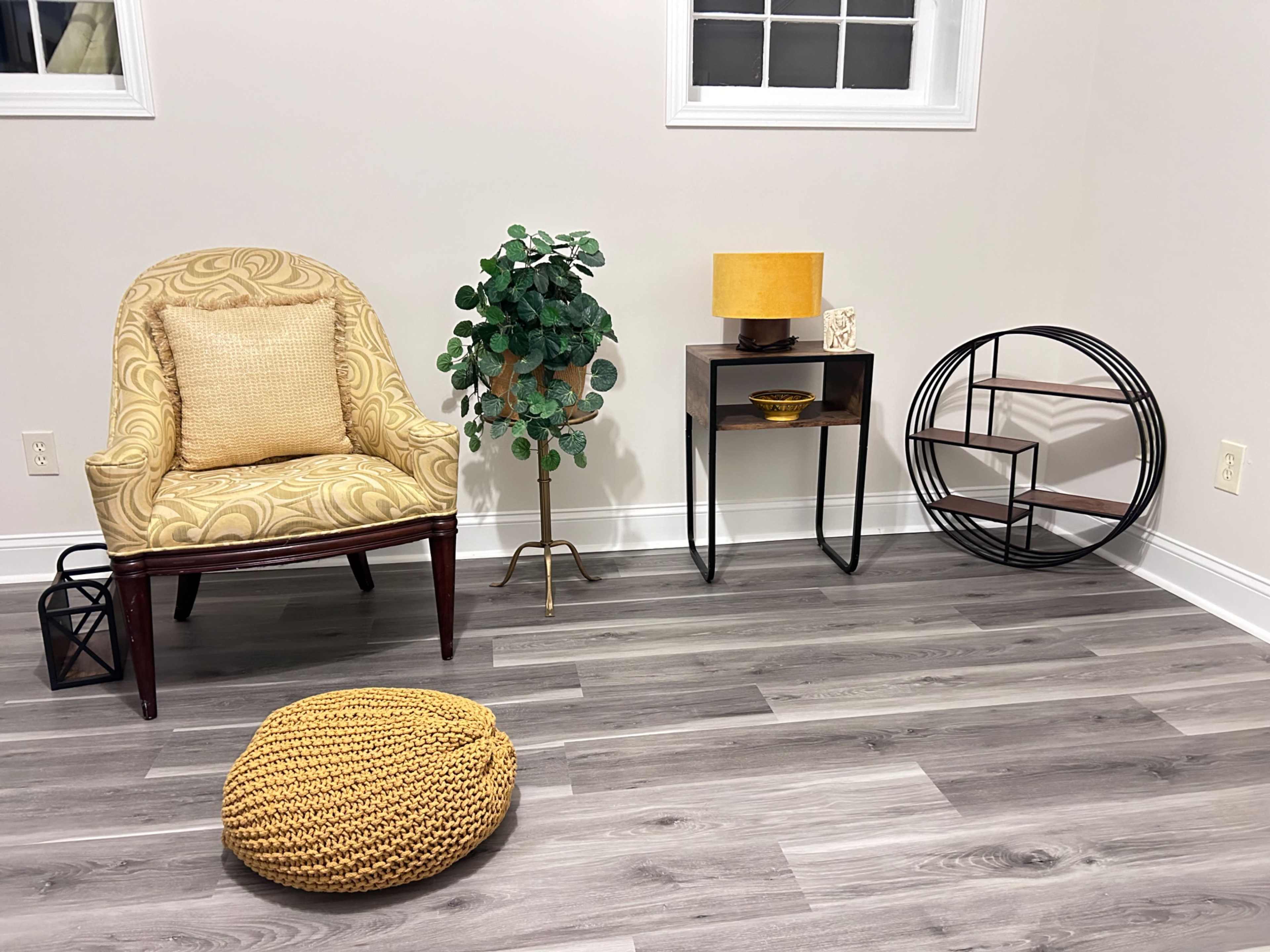 The image shows a minimalist room with a patterned chair, a decorative plant, a table lamp, a small side table, and a round pouf on wooden flooring.