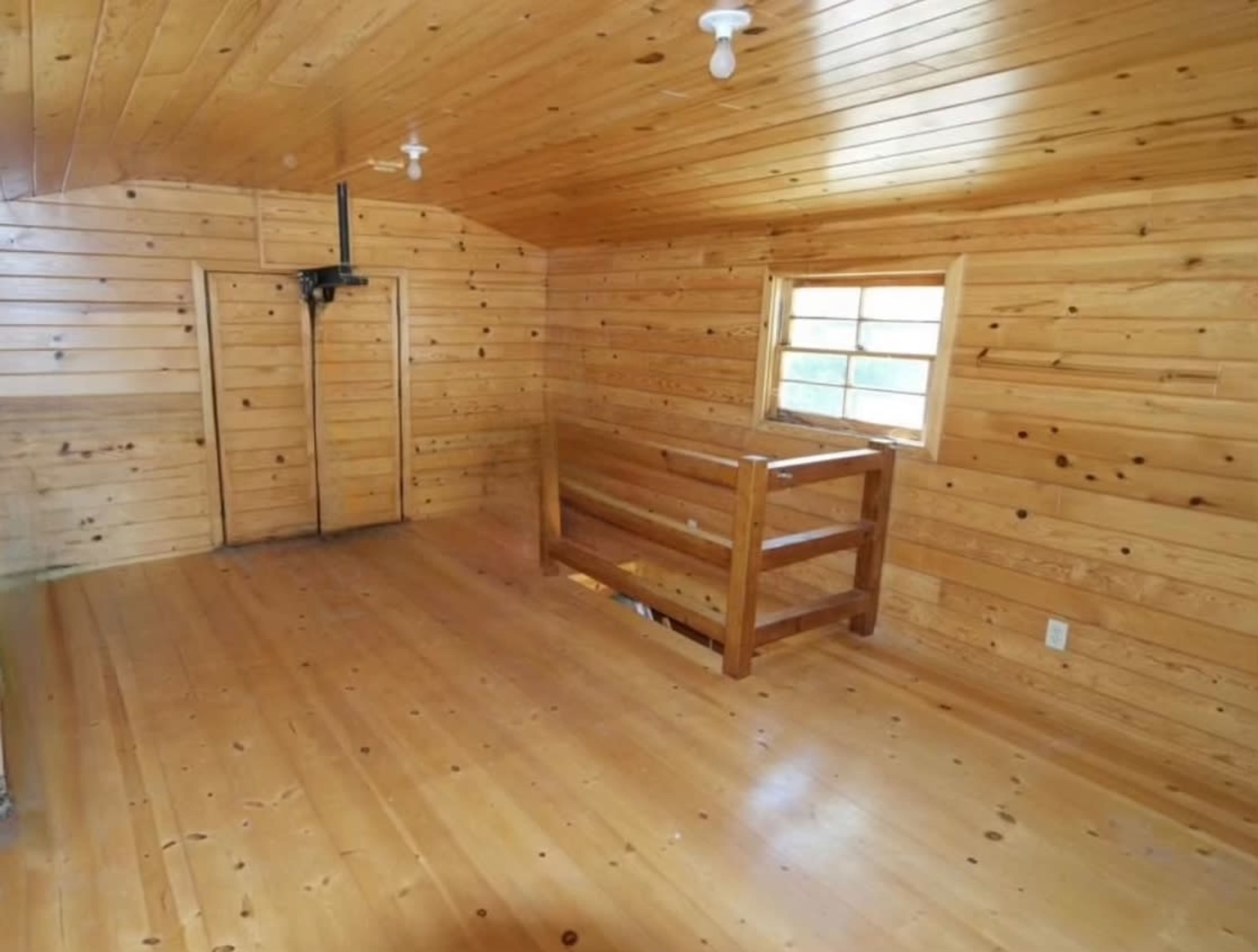 An empty wooden room with pine paneling on the walls and ceiling, a window, and a wooden railing near the back.