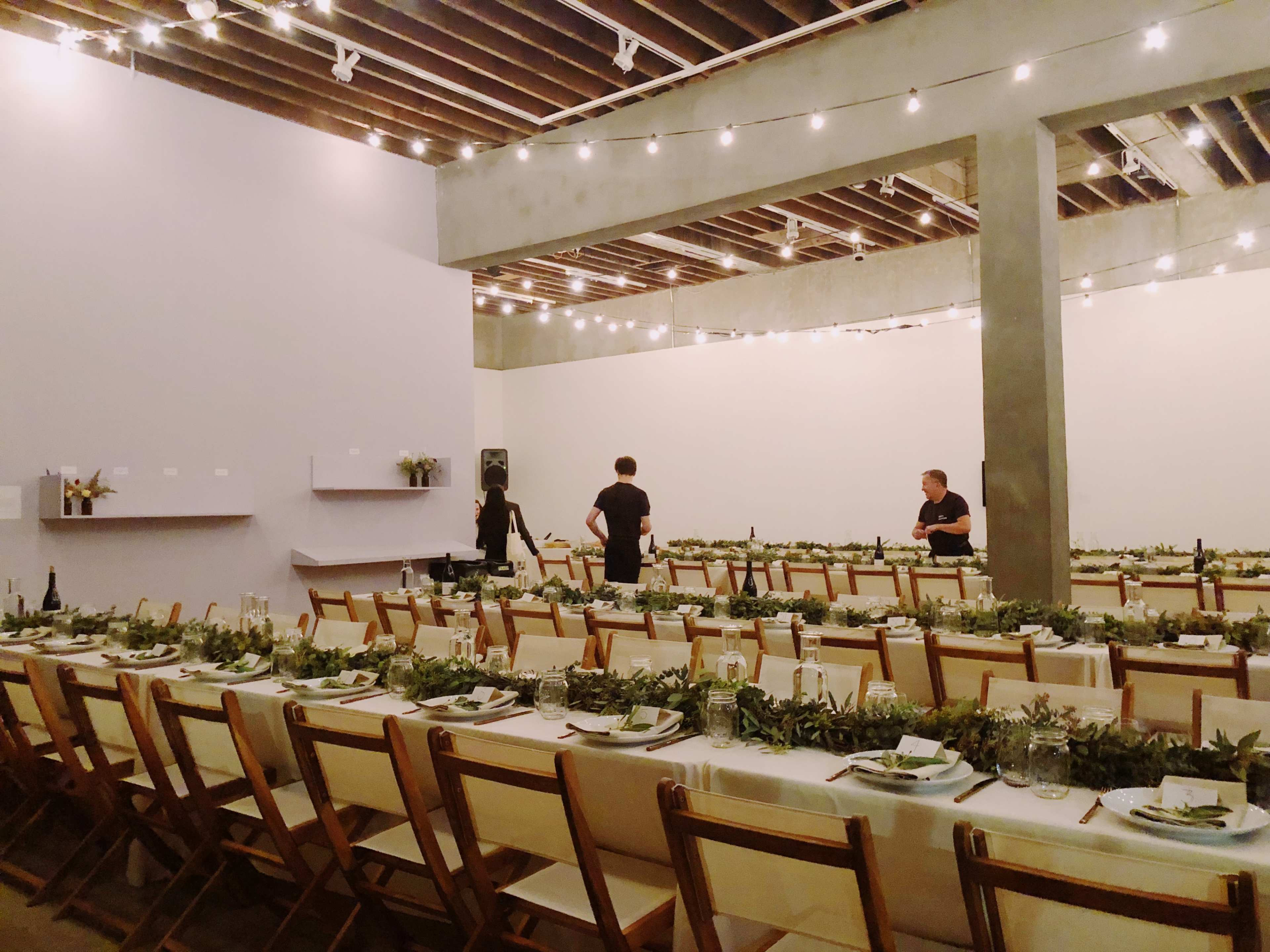 A spacious dining area is set with long tables adorned with greenery and table settings, while two people prepare the space under string lights.