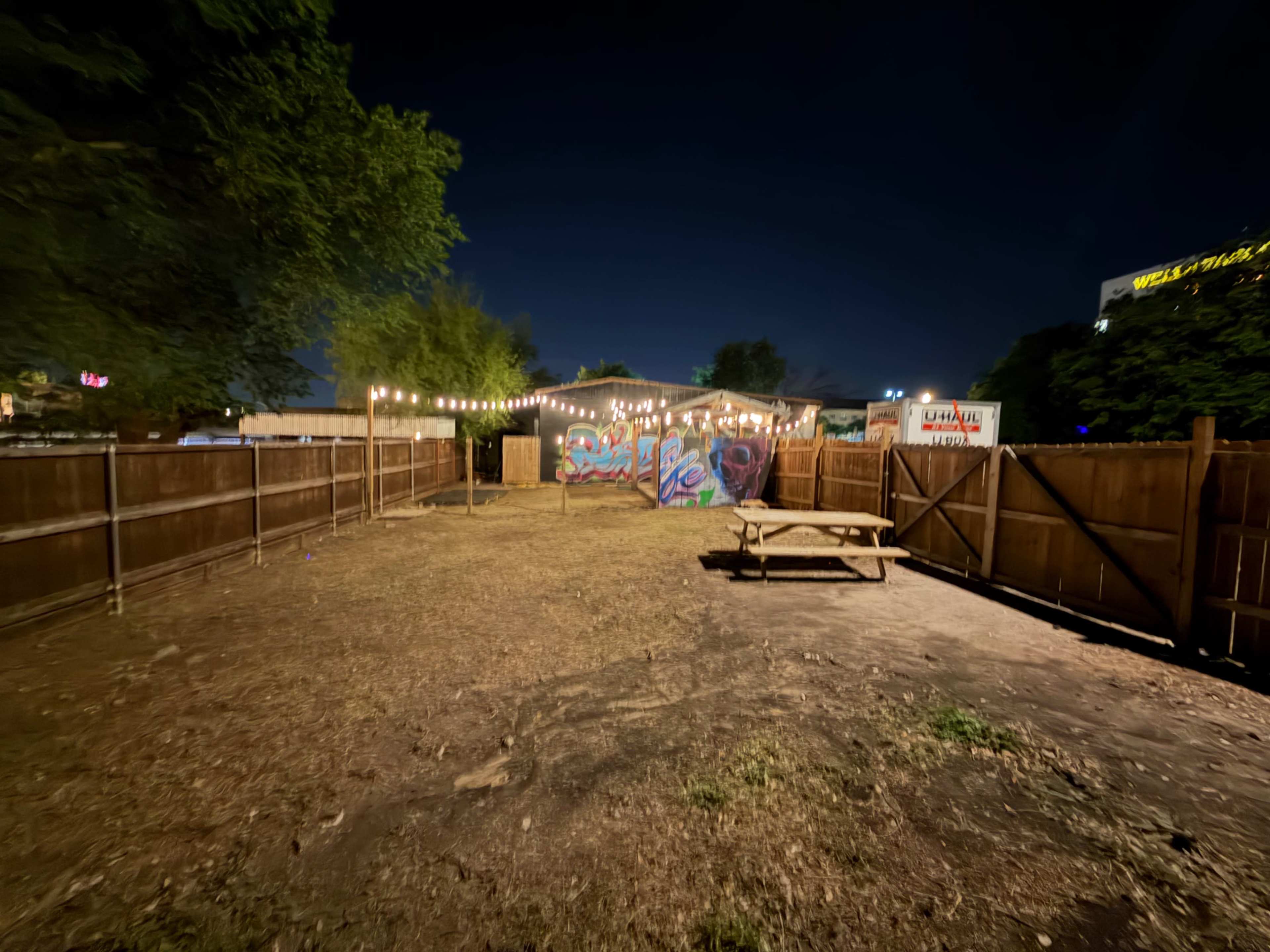 The image shows an outdoor space at night featuring a fenced area with a wooden picnic table and a wall adorned with colorful graffiti.