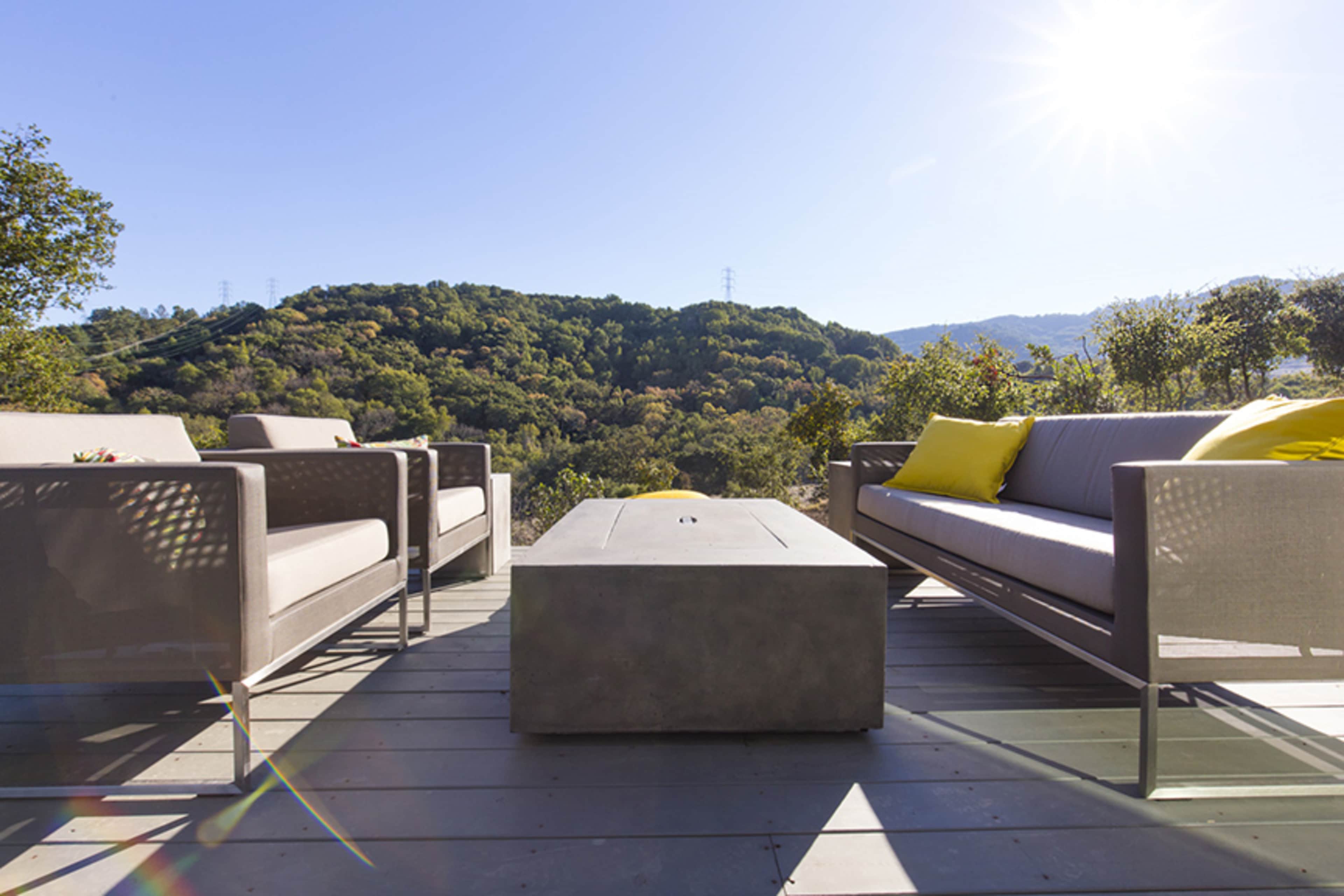 A modern outdoor seating area features two sofas and a rectangular coffee table, surrounded by lush green hills and clear blue skies.