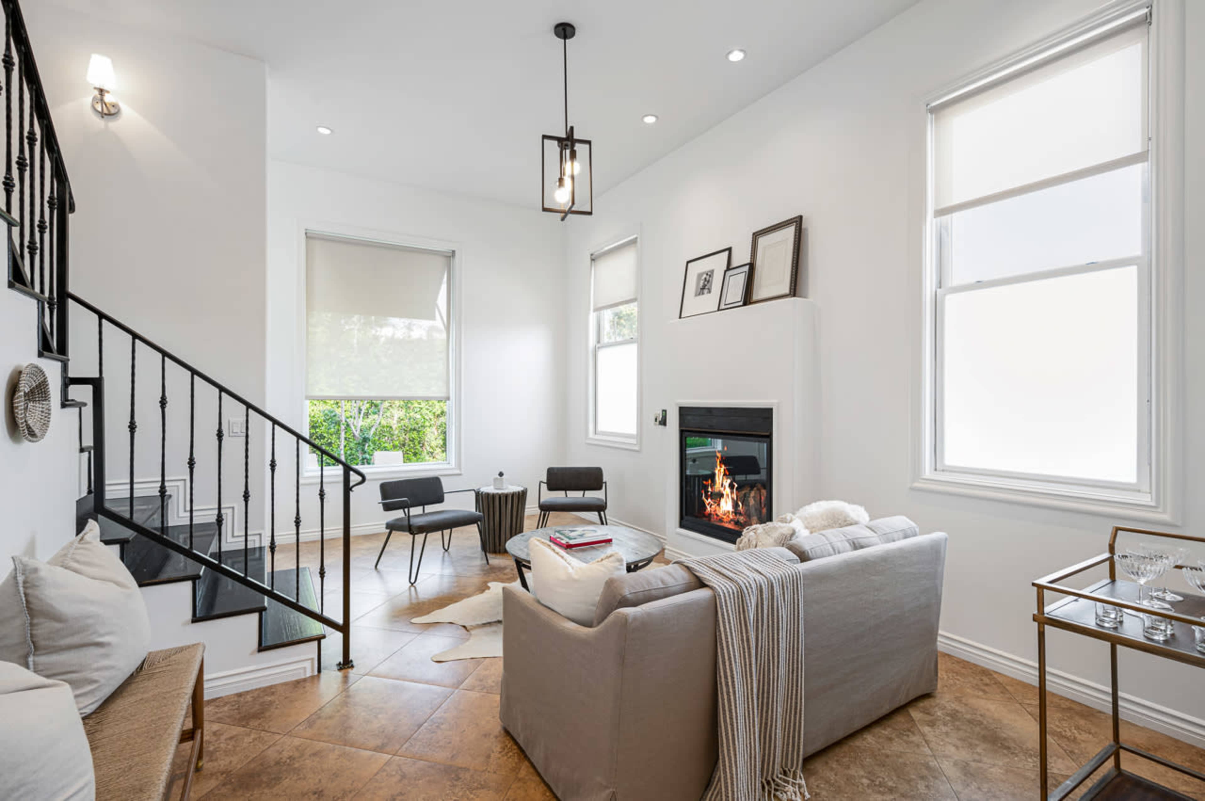 A modern living room features a gray sofa, a fireplace, and large windows, with a staircase in the background.