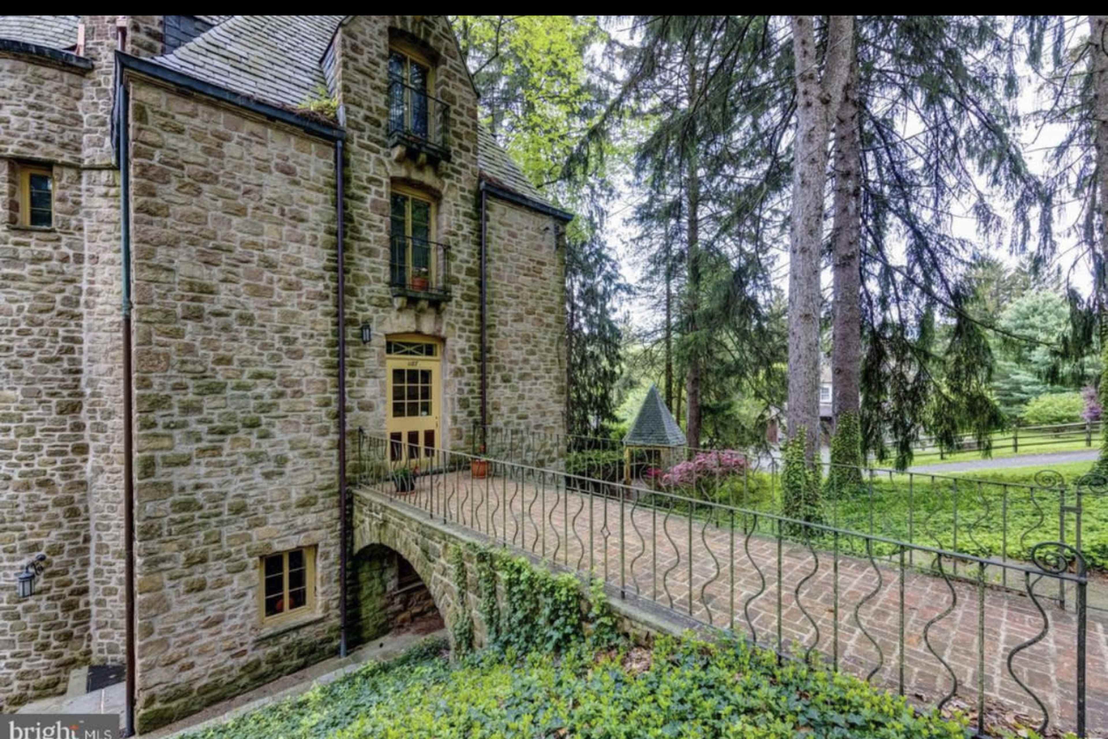 A stone house with an arched bridge leads to its entrance, surrounded by tall trees and green landscaping.