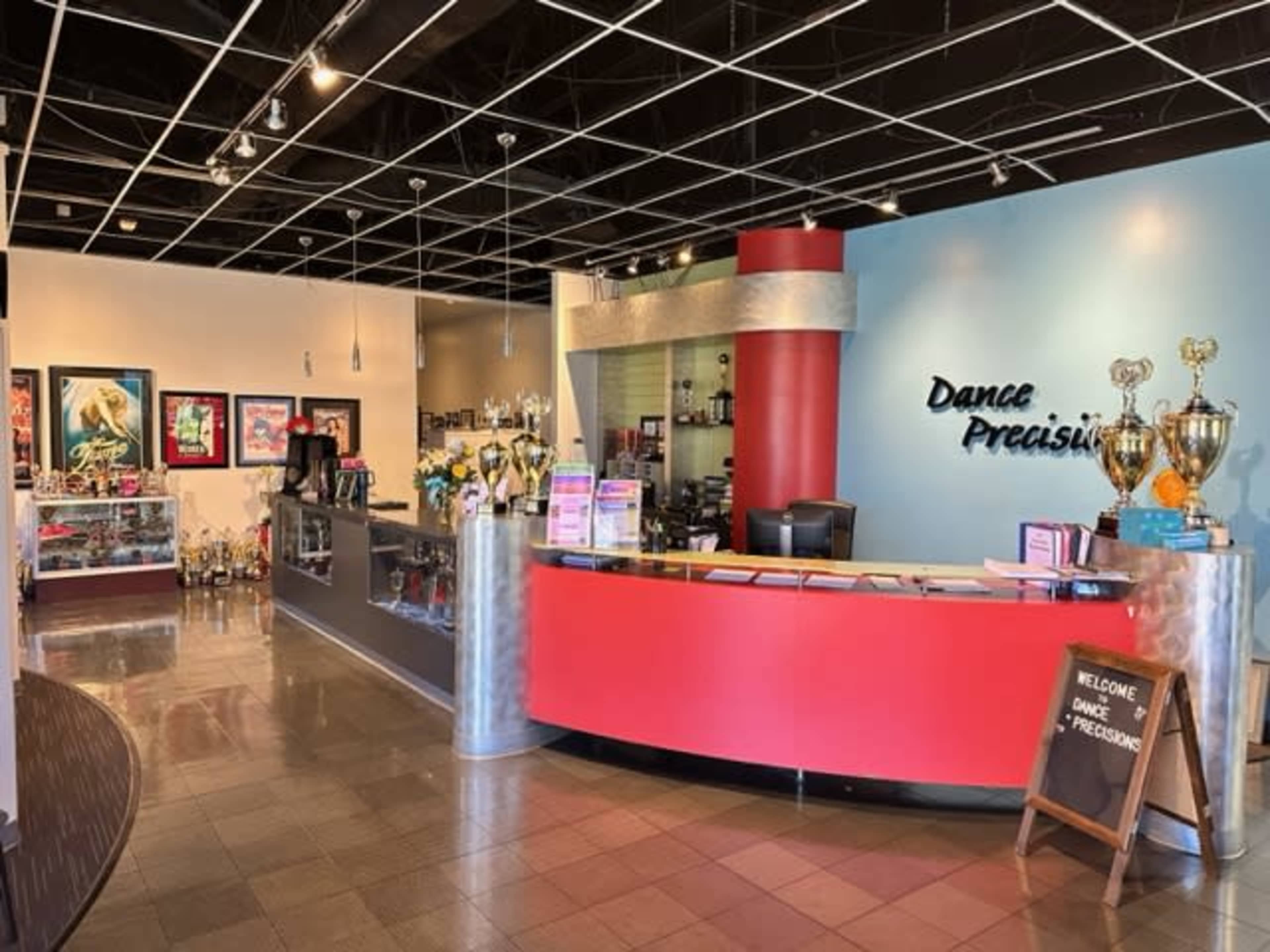 The interior of a dance studio with a reception area featuring a red desk, trophies on display, and framed posters on the walls.
