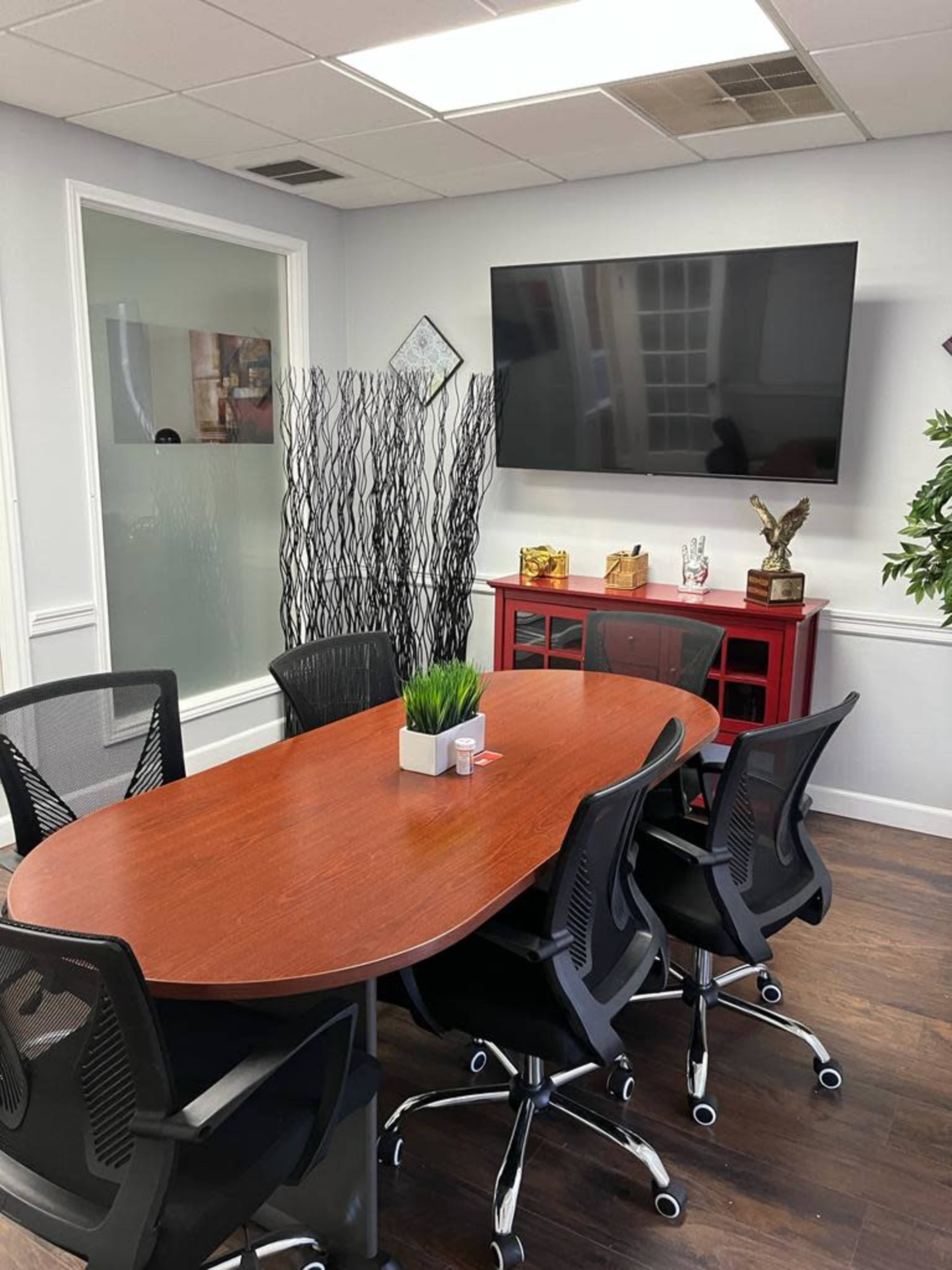 A modern conference room featuring a wooden oval table surrounded by black mesh chairs, a large flat-screen TV mounted on the wall, and a red cabinet displaying decorative items.