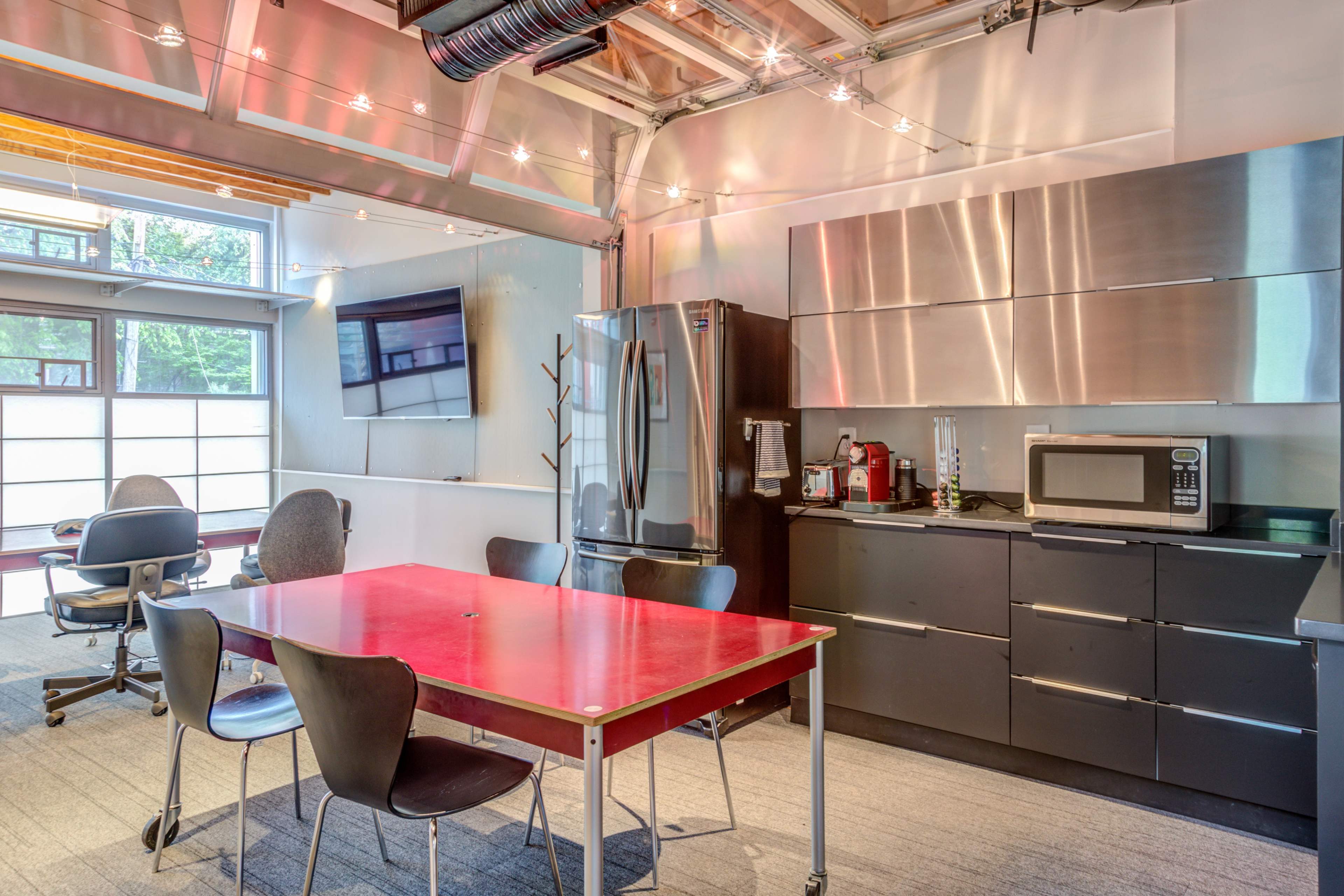 A modern office kitchen with a red table, black cabinets, stainless steel appliances, and seating arranged nearby.