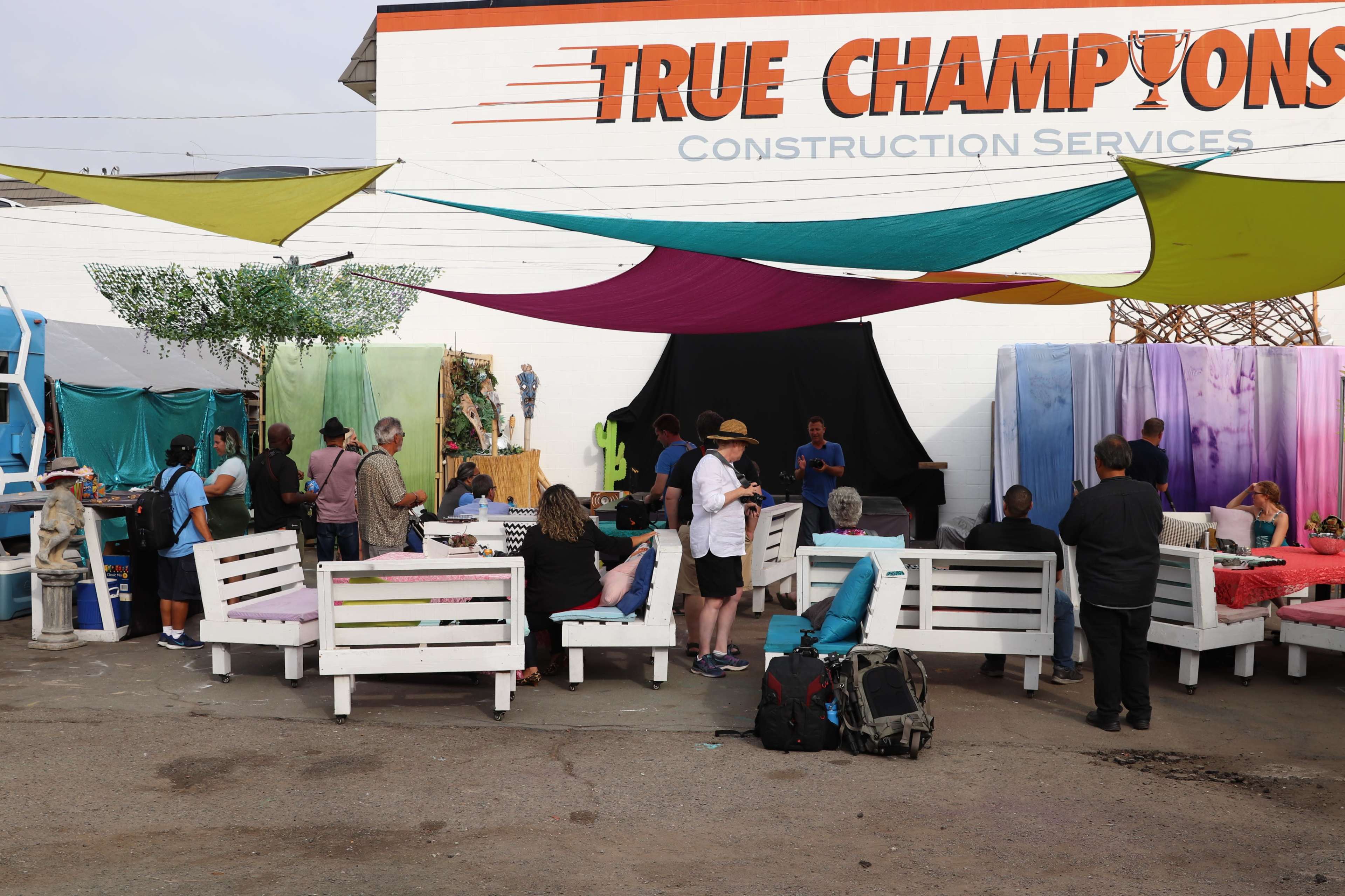A group of people gathers in an outdoor space with colorful fabric awnings and white wooden benches in front of a wall advertising "True Champions Construction Services."