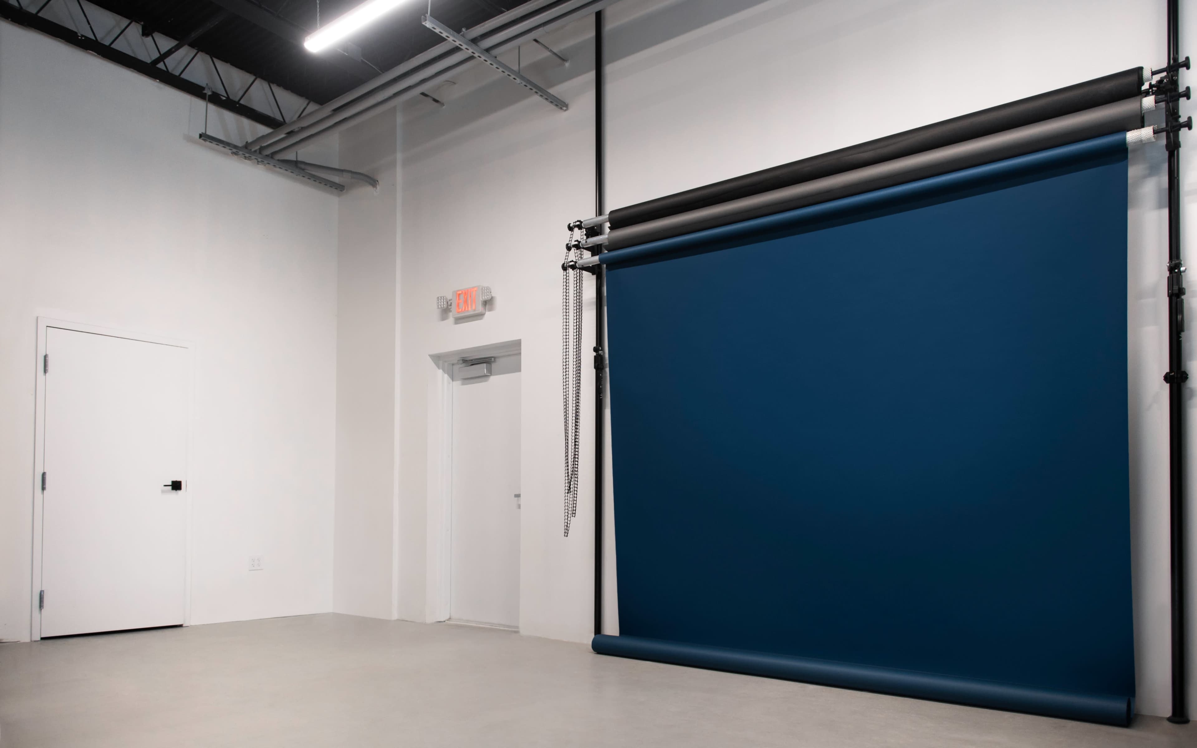 The image shows a minimalist studio space with a dark blue backdrop and a white wall, featuring an exit door.
