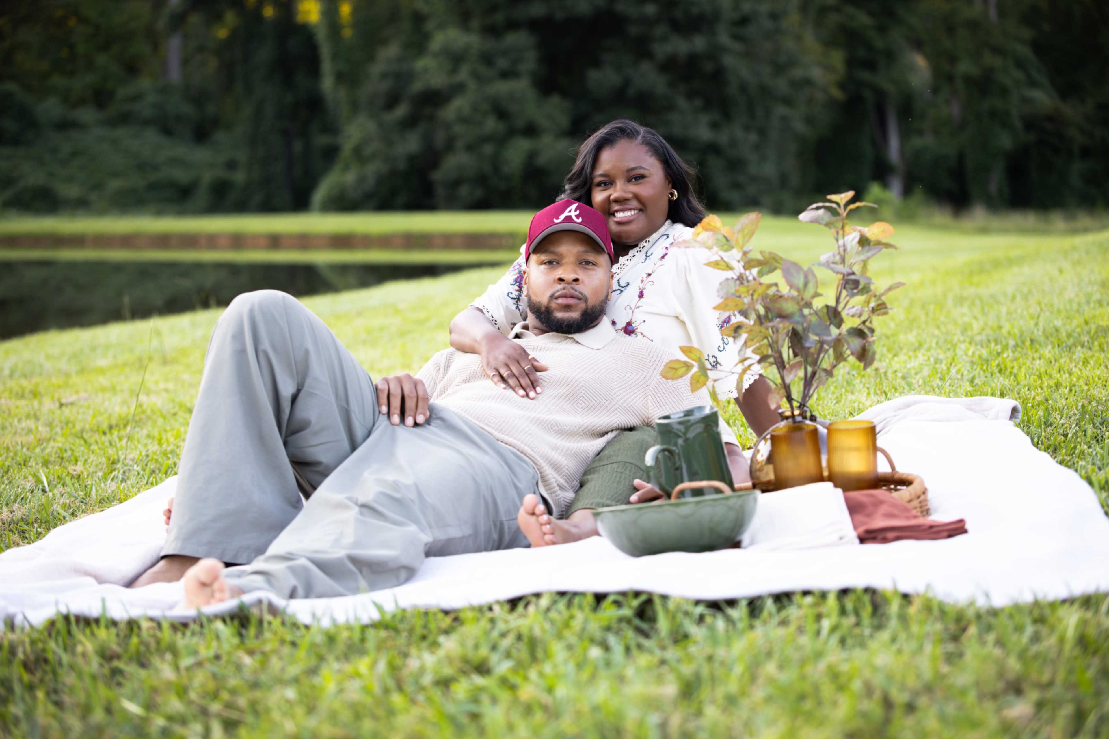 A couple relaxes on a white blanket beside a pond, surrounded by plants and decorative items.