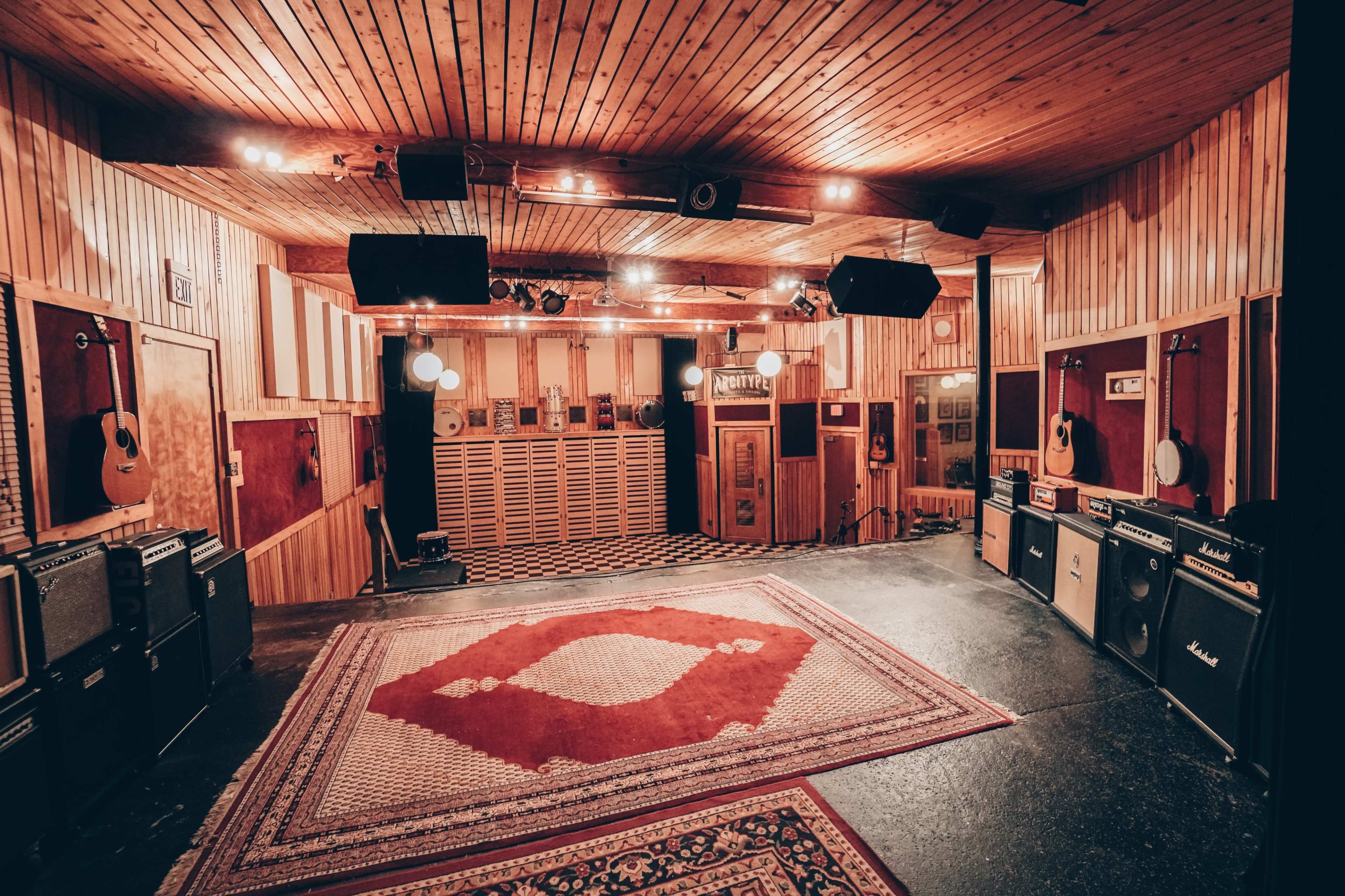 A spacious recording studio with wooden walls, a patterned rug on the floor, and musical instruments including guitars and amplifiers positioned around the room.