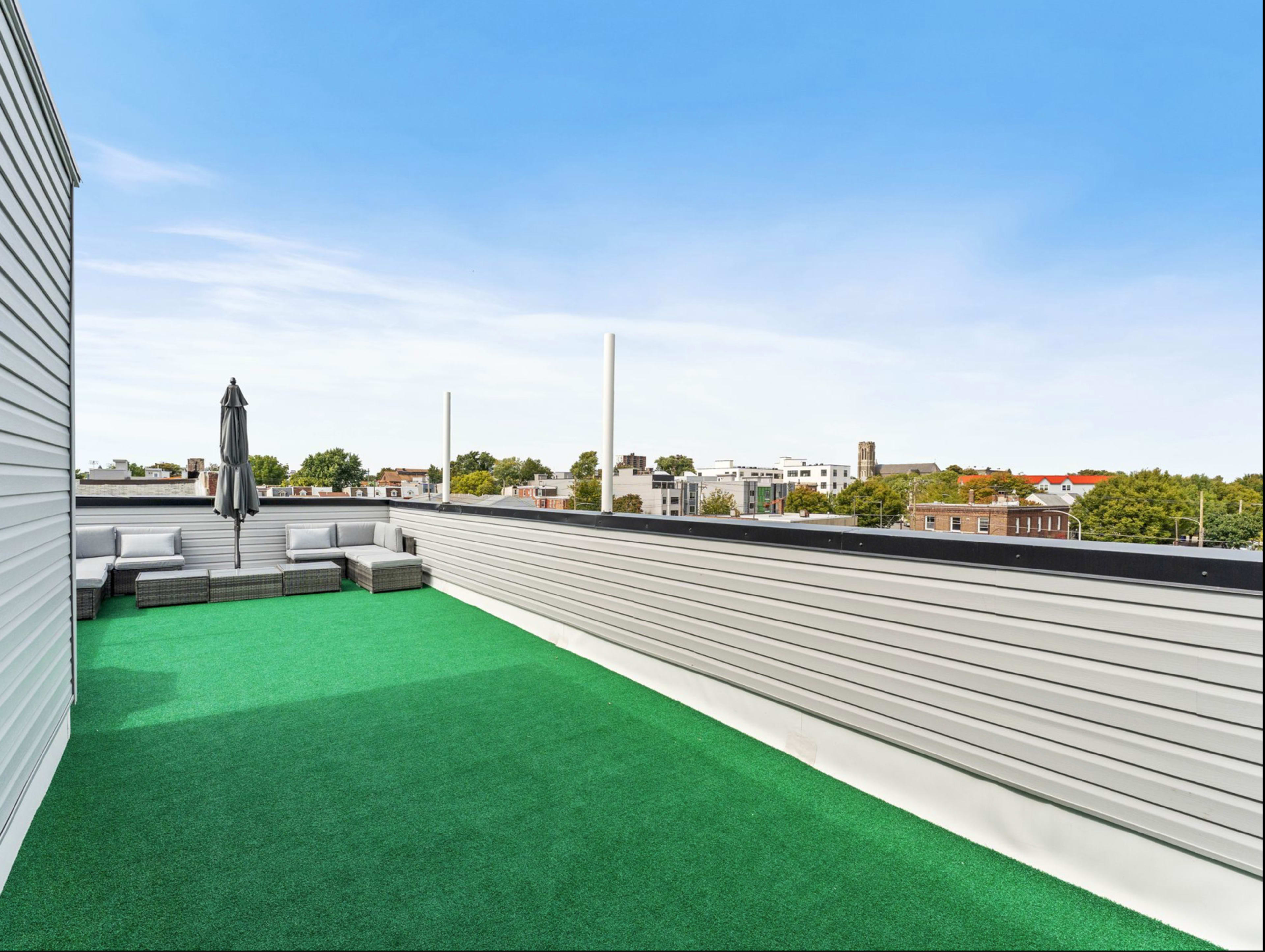 The image shows a rooftop terrace with artificial grass, a seating area, and a clear blue sky in the background.