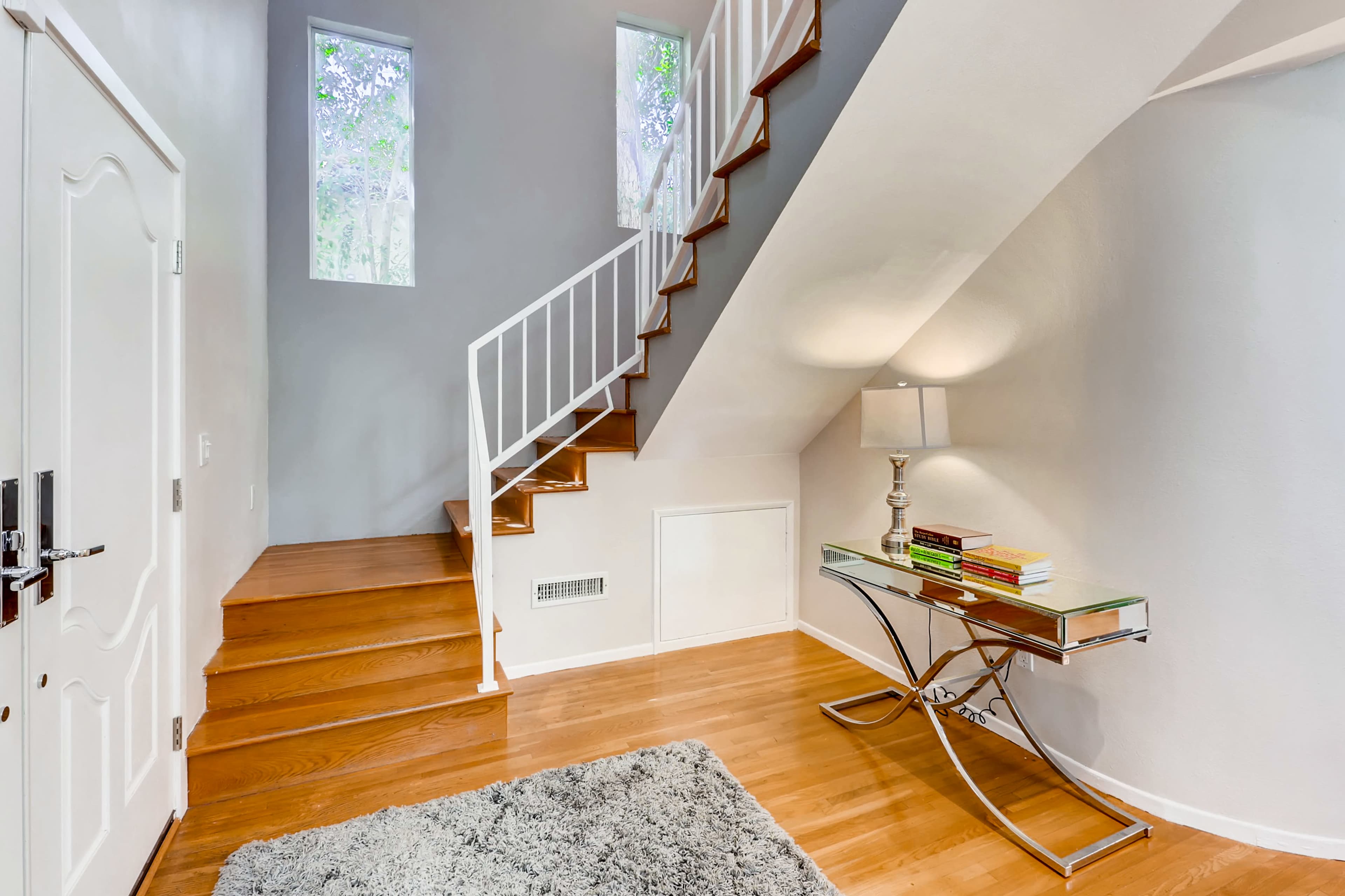 A bright entryway with a staircase leading to an upper level, wooden floors, and a glass table with books and a lamp.