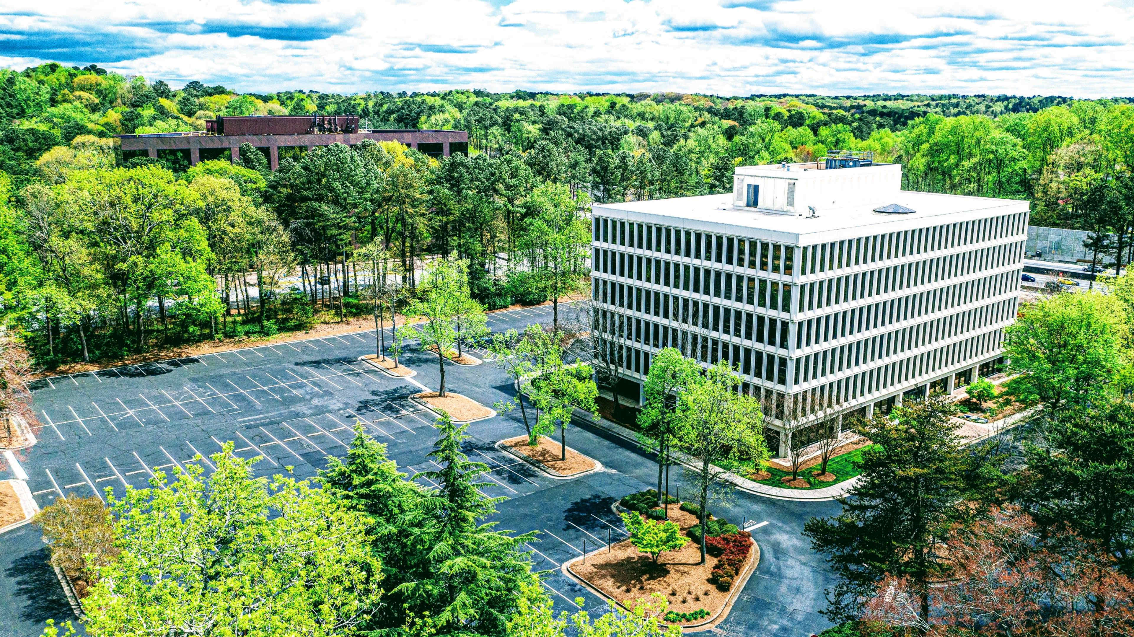 A modern office building stands next to a largely empty parking lot surrounded by greenery and trees under a partly cloudy sky.