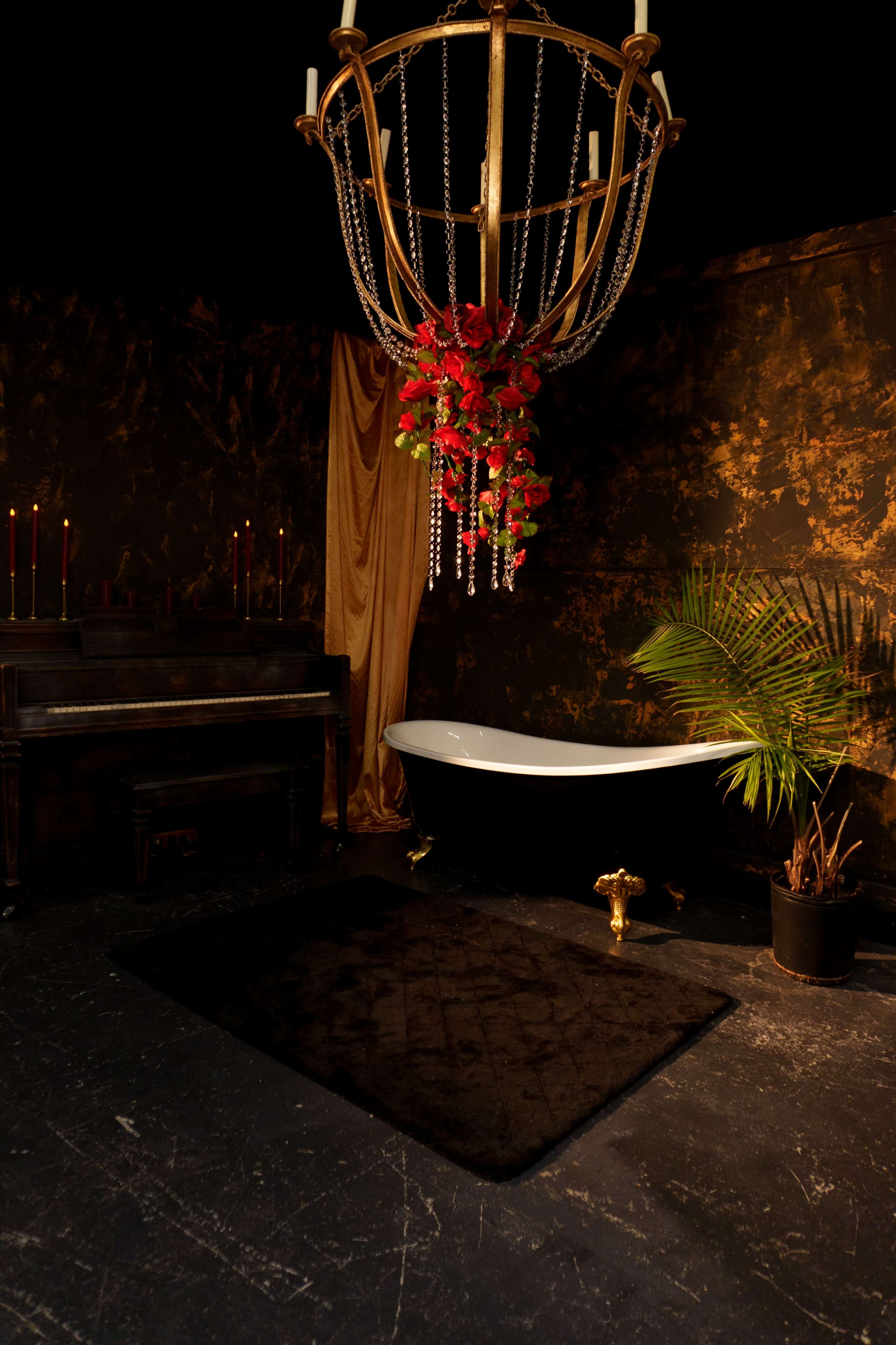 The image features a vintage bathroom with a freestanding white bathtub, a large chandelier adorned with flowers, a dark rug, and a piano in a dimly lit, ornate setting.
