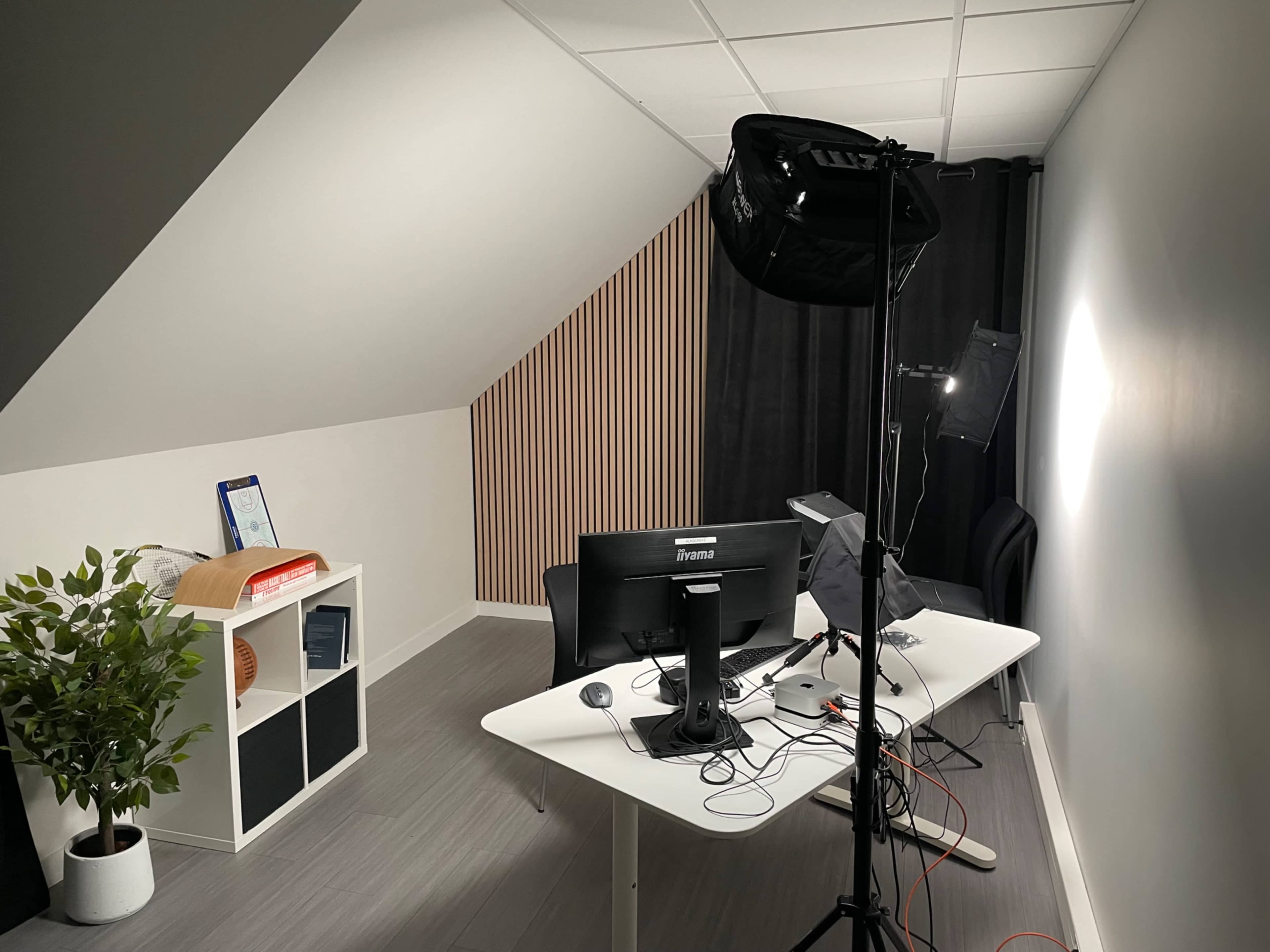 The image shows a modern, minimalistic workspace with a desk, computer setup, and photographic lighting equipment in a well-lit room.