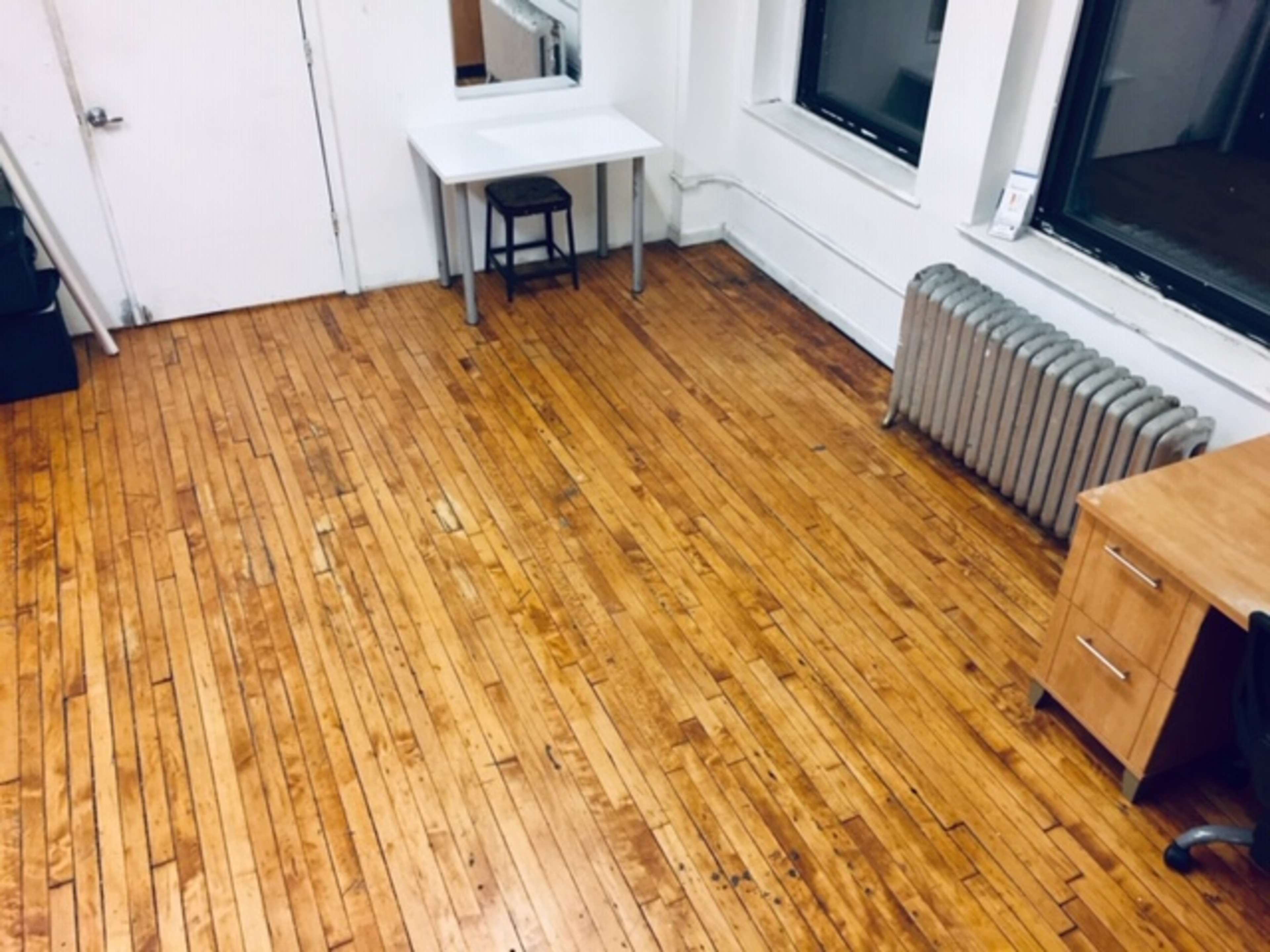A large room with wooden flooring, a table and chair, a radiator, and a door leading to an exterior.