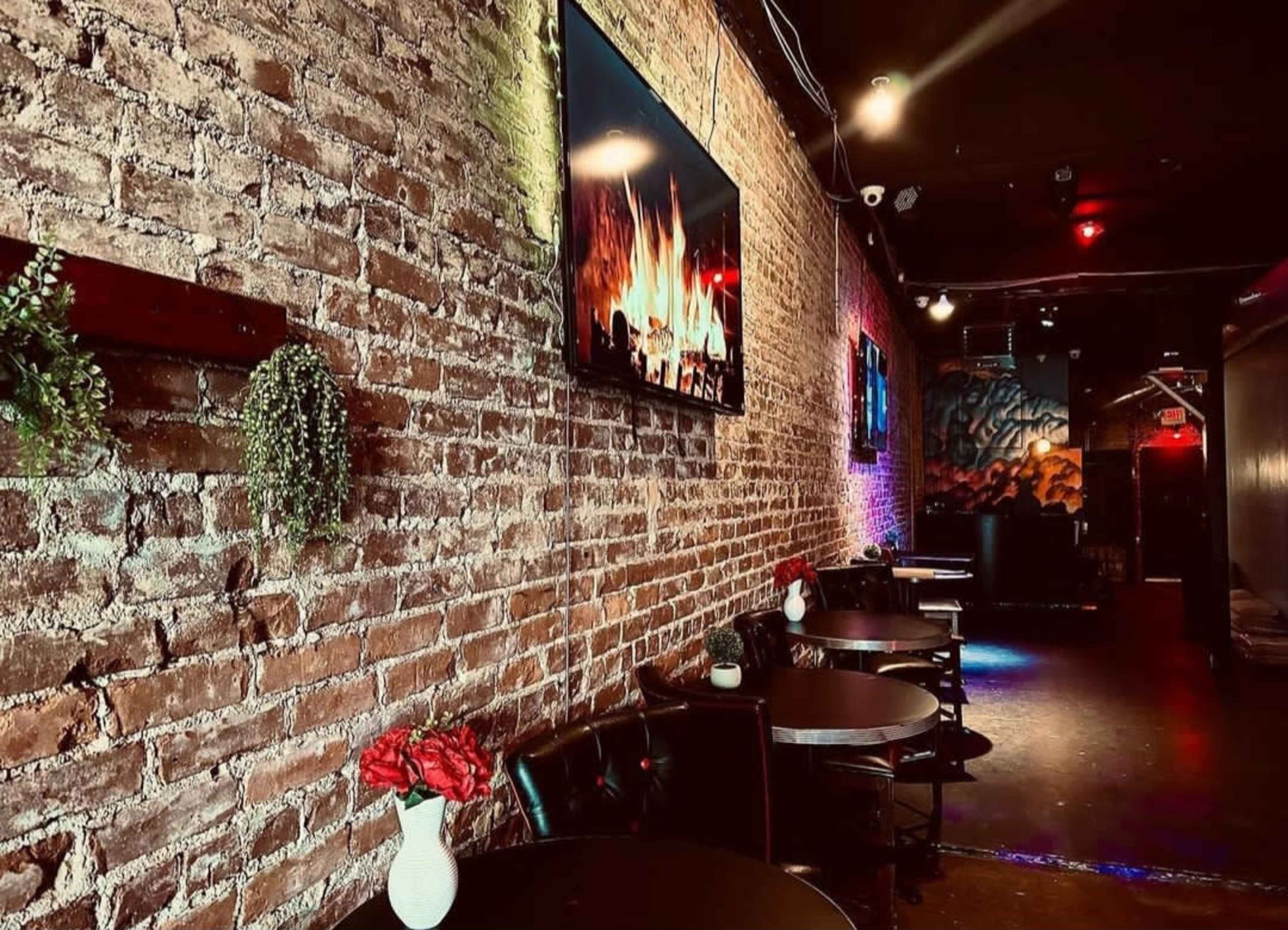 The image shows a dimly lit restaurant with exposed brick walls, a television displaying a fireplace, and several tables adorned with small vases of red flowers.