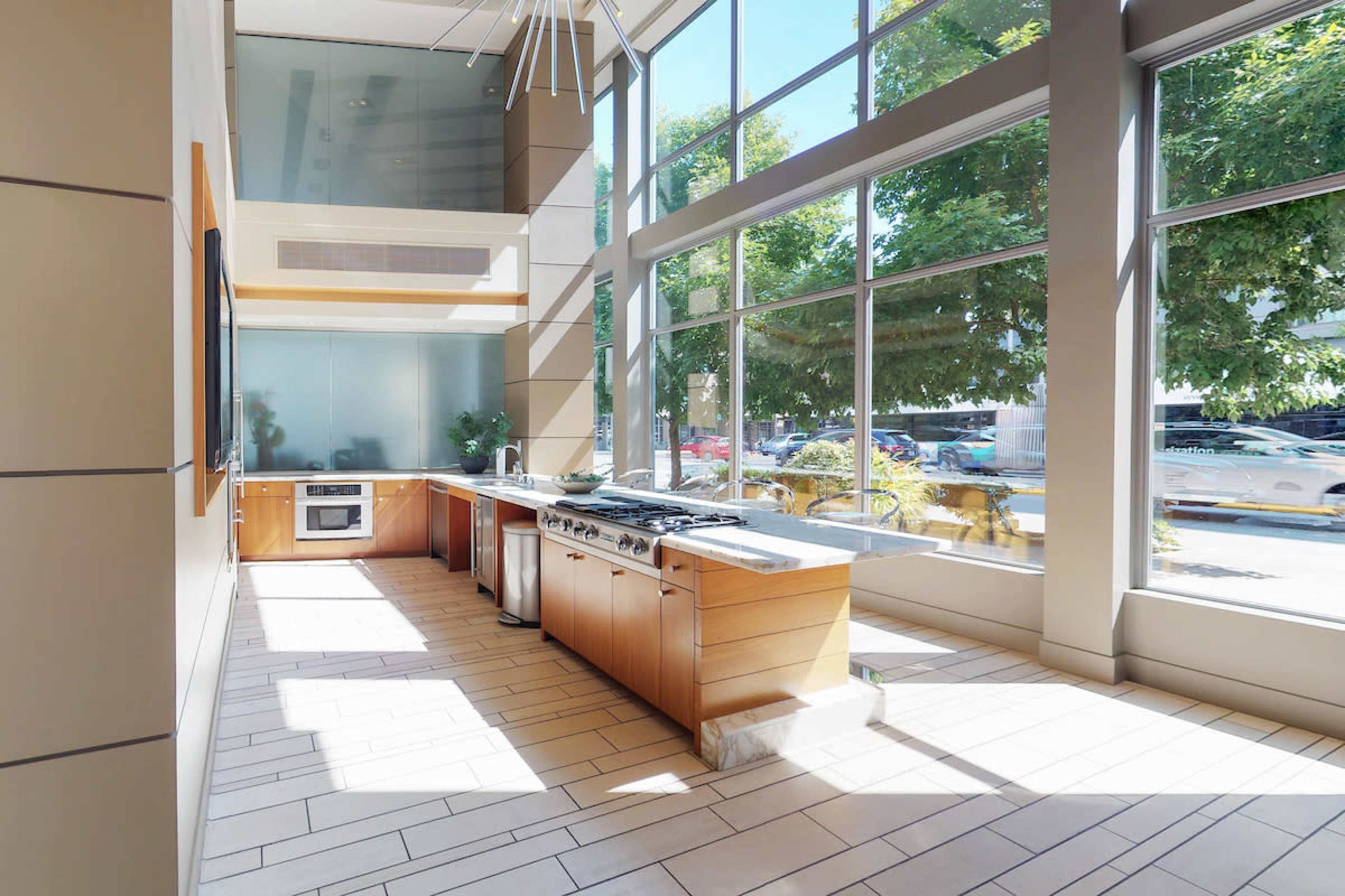 The image shows a modern kitchen space with large windows, featuring wooden cabinetry, a stainless steel oven, and a gas stovetop.