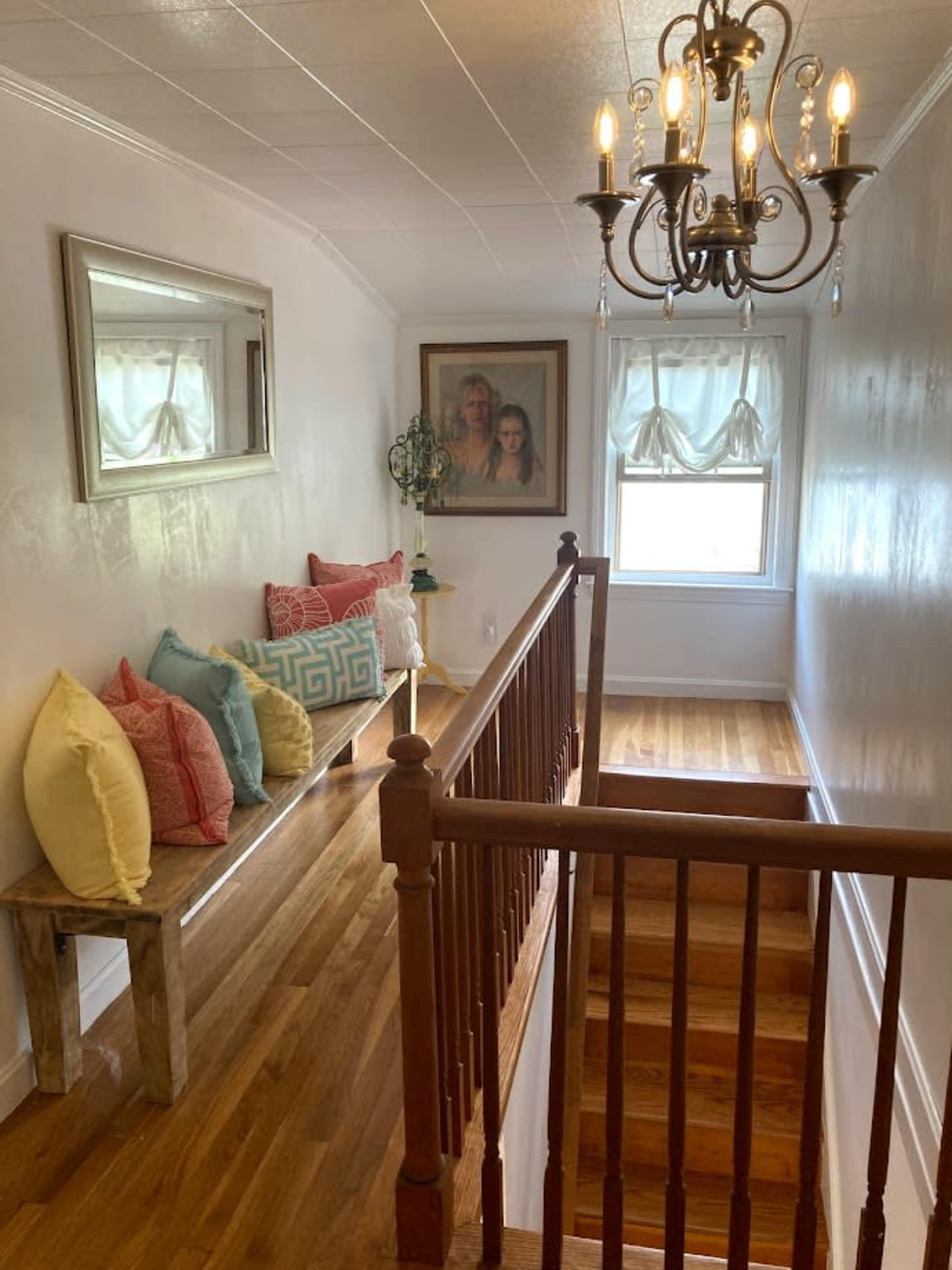 A well-lit hallway with a wooden railing, a small table with colorful pillows, a framed picture on the wall, and a chandelier overhead.