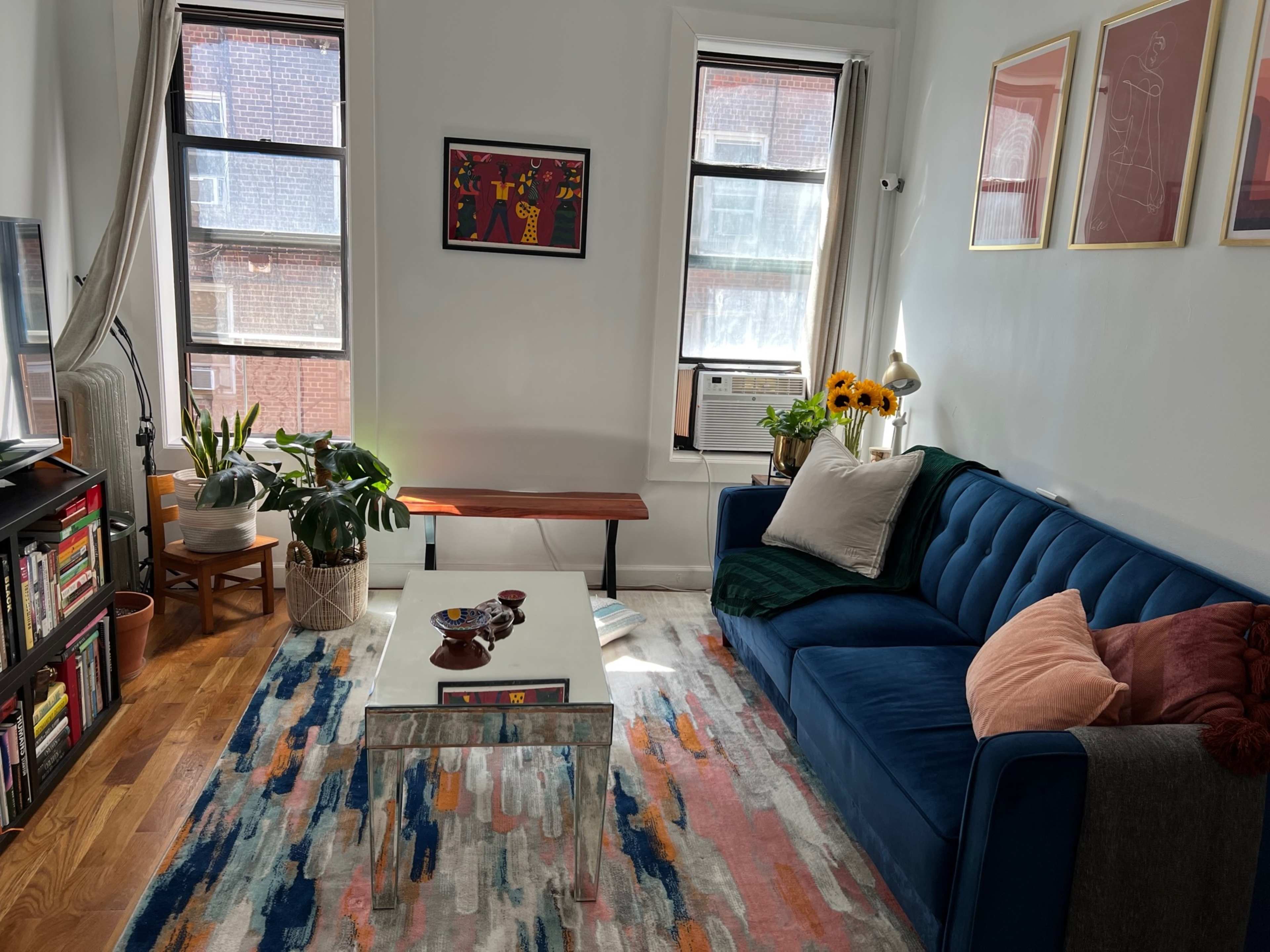 The living room features a blue sofa, a glass coffee table, a large area rug, a few plants, and a mix of artwork on the walls.