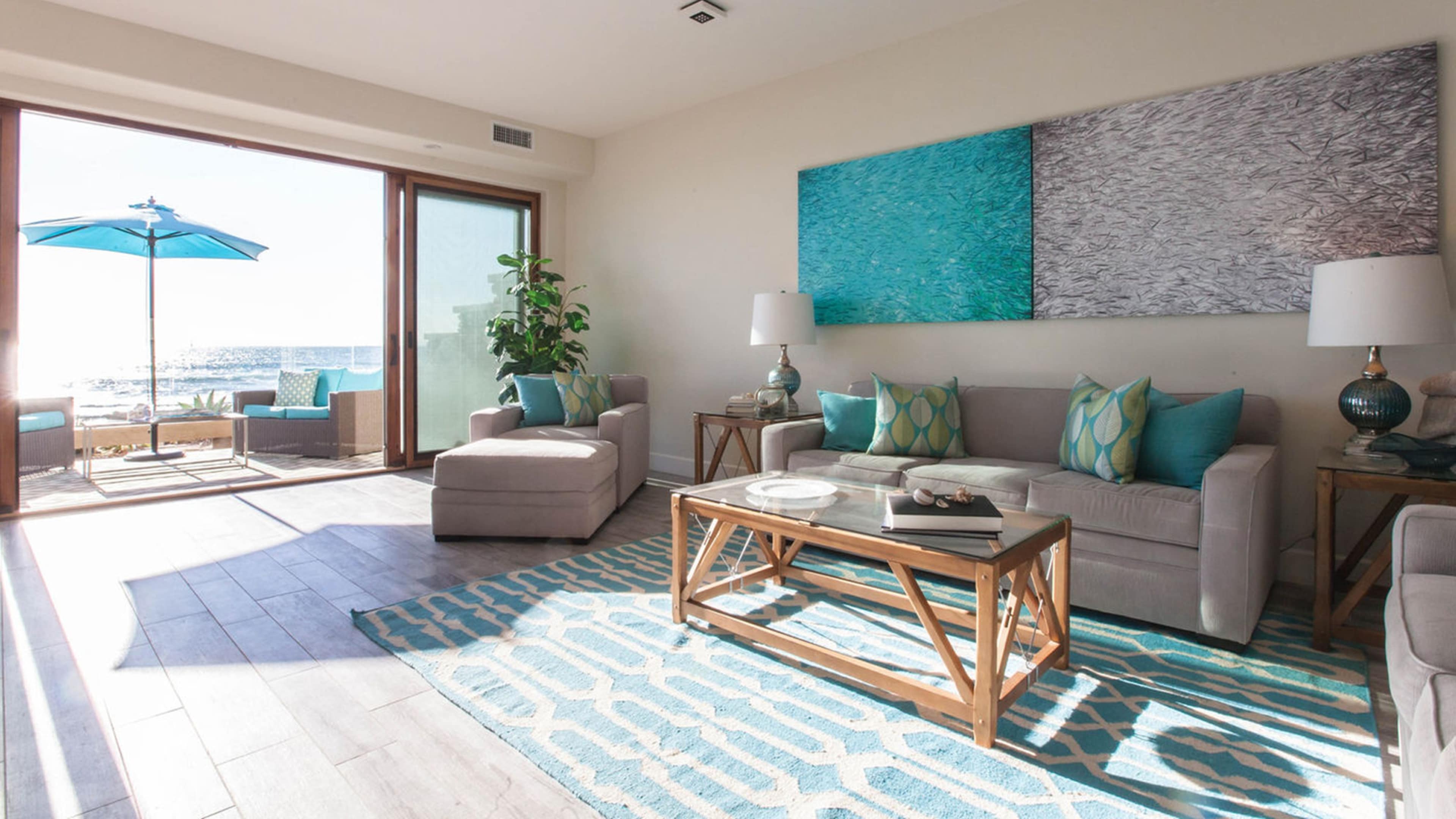 The living room features large windows with ocean views, a light-colored sofa and chairs, a wooden coffee table, and blue accent decor.