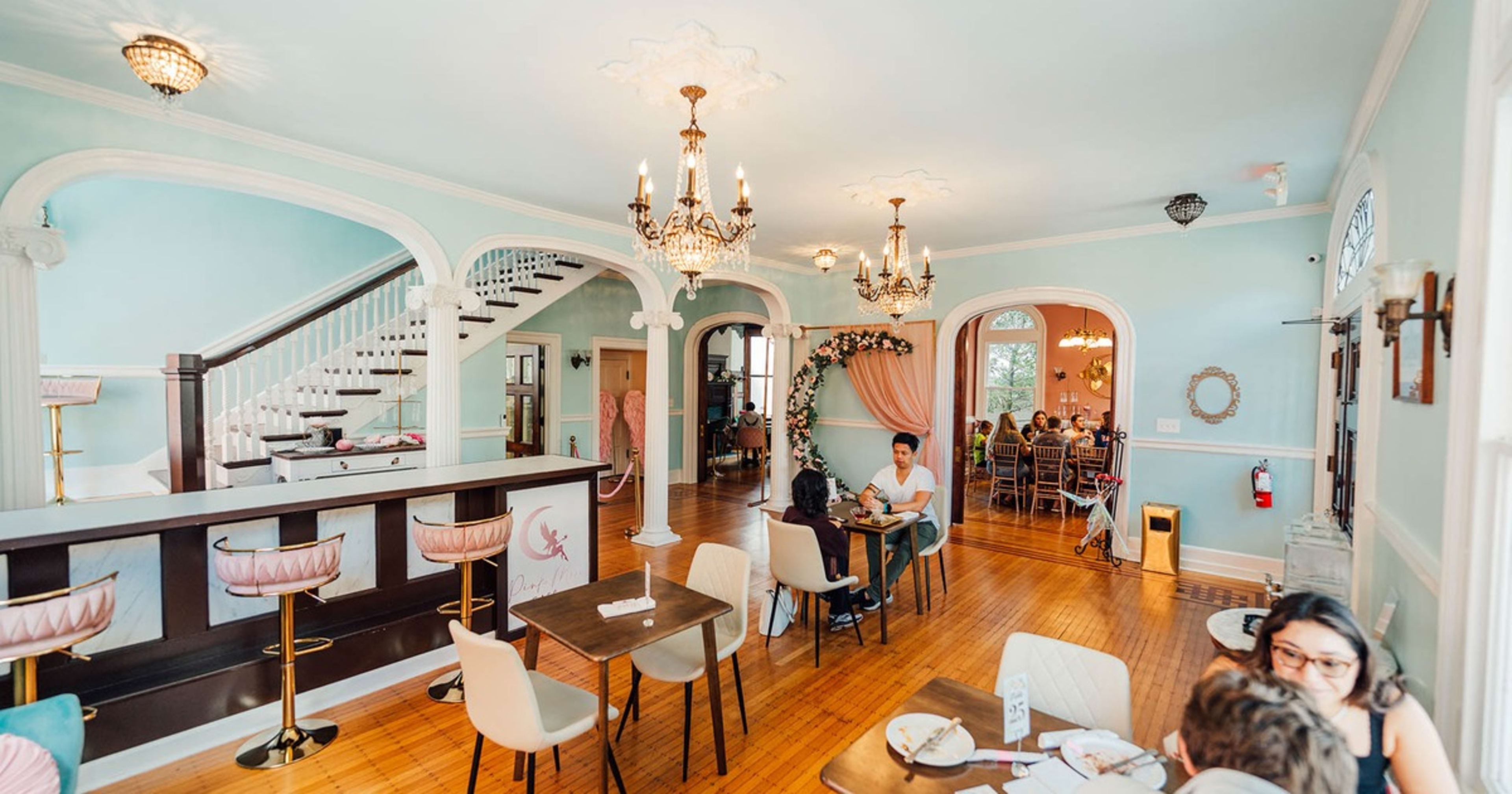 A spacious, pastel-colored café featuring elegant chandeliers, a staircase, and various seating options.