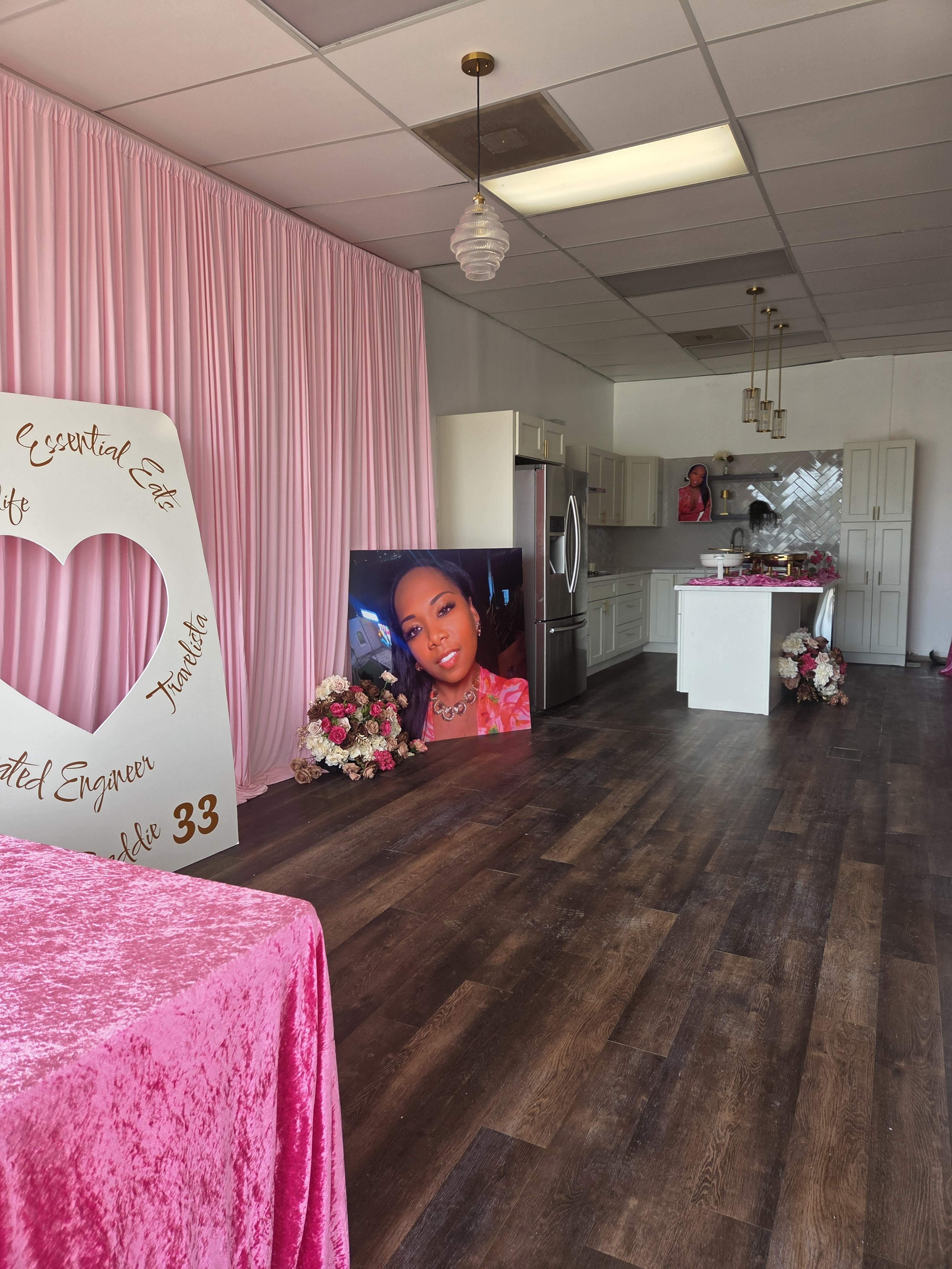 The image shows a brightly lit room with pink curtains, a large framed photo of a woman, and floral decorations around a display.