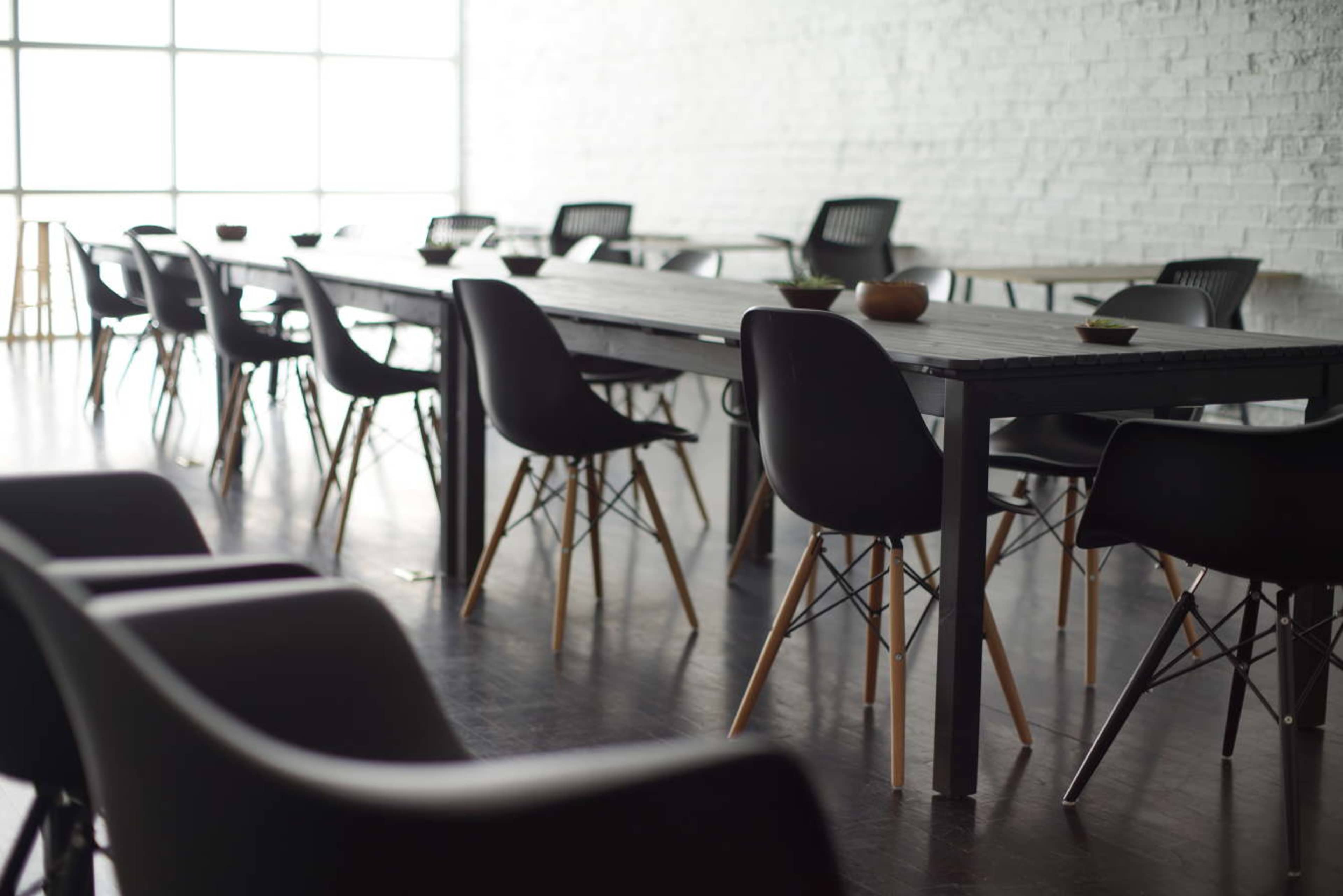 A spacious room features a long wooden table surrounded by sleek black chairs on a polished floor, with large windows letting in natural light.