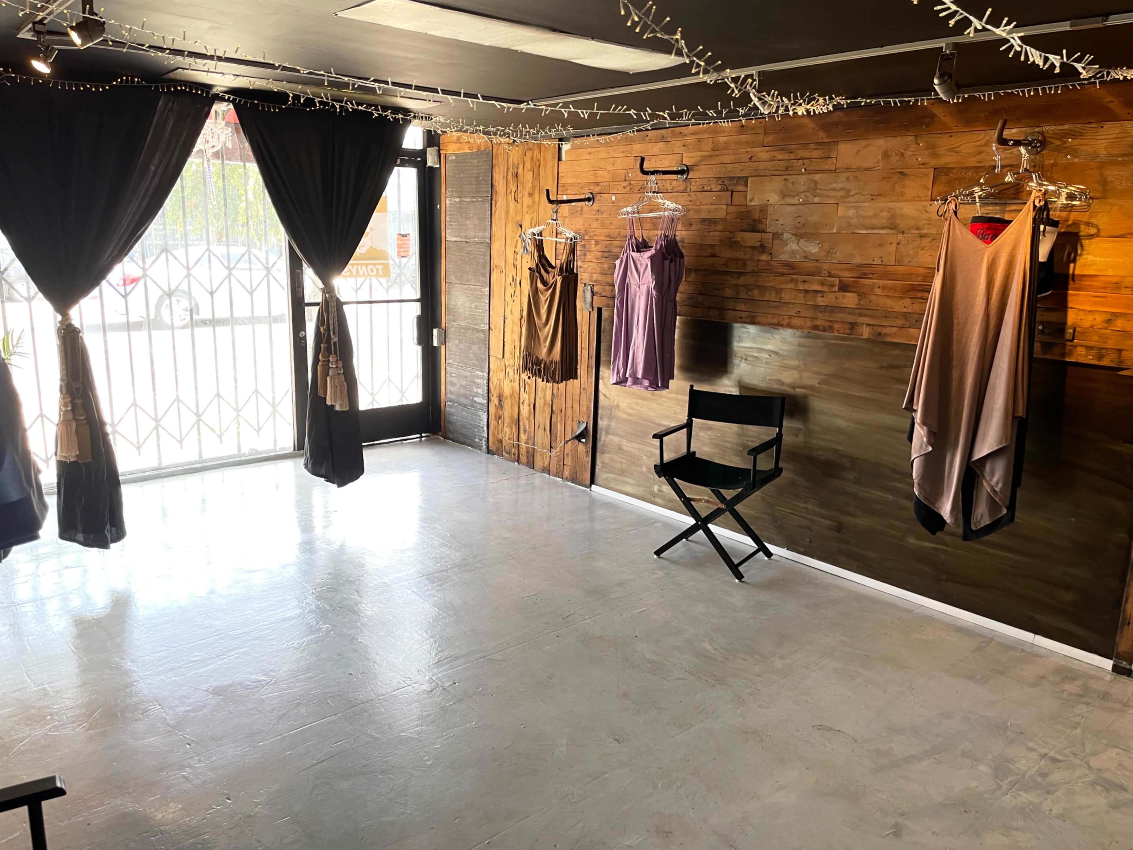 The image shows an empty clothing boutique with wooden walls, hanging dresses, and a director's chair near the entrance.