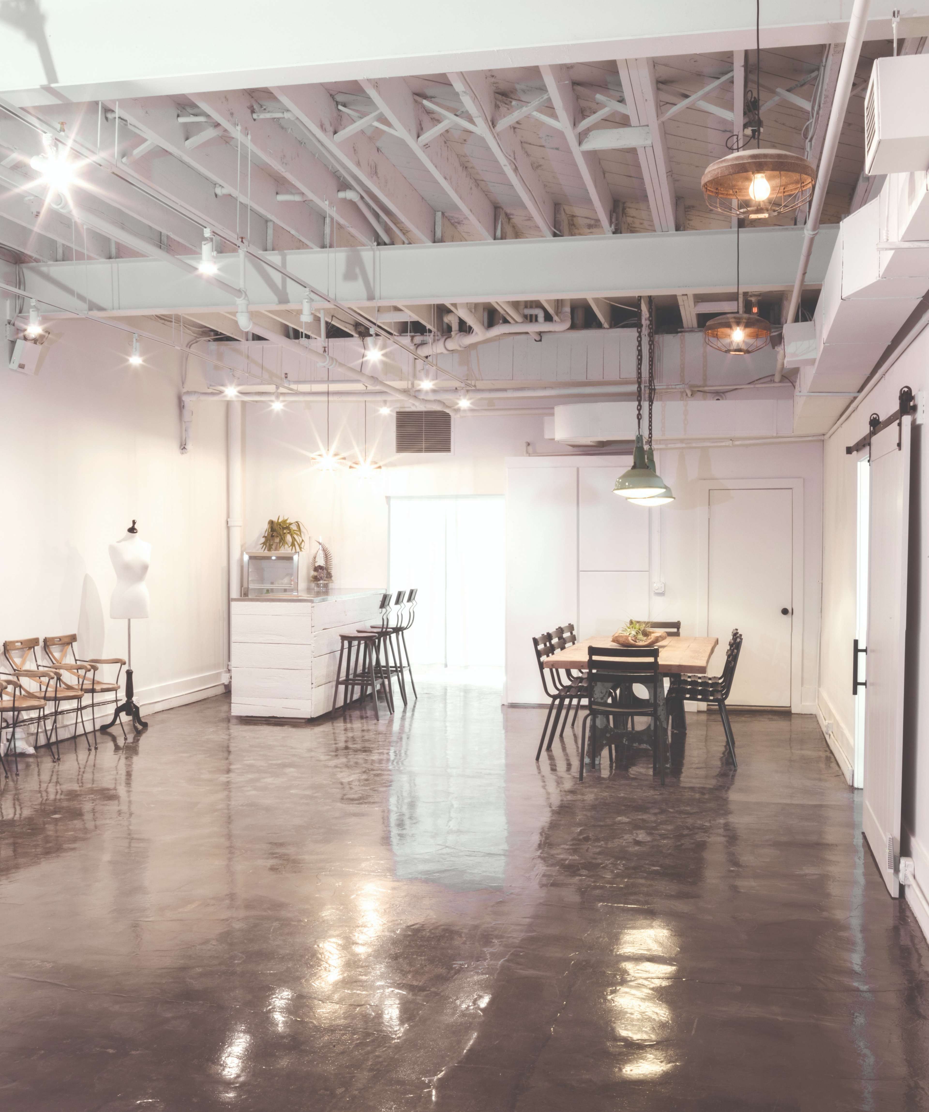 The image shows a spacious, well-lit room with a polished concrete floor, featuring a dining table and several chairs, along with a mannequin and a white dresser.