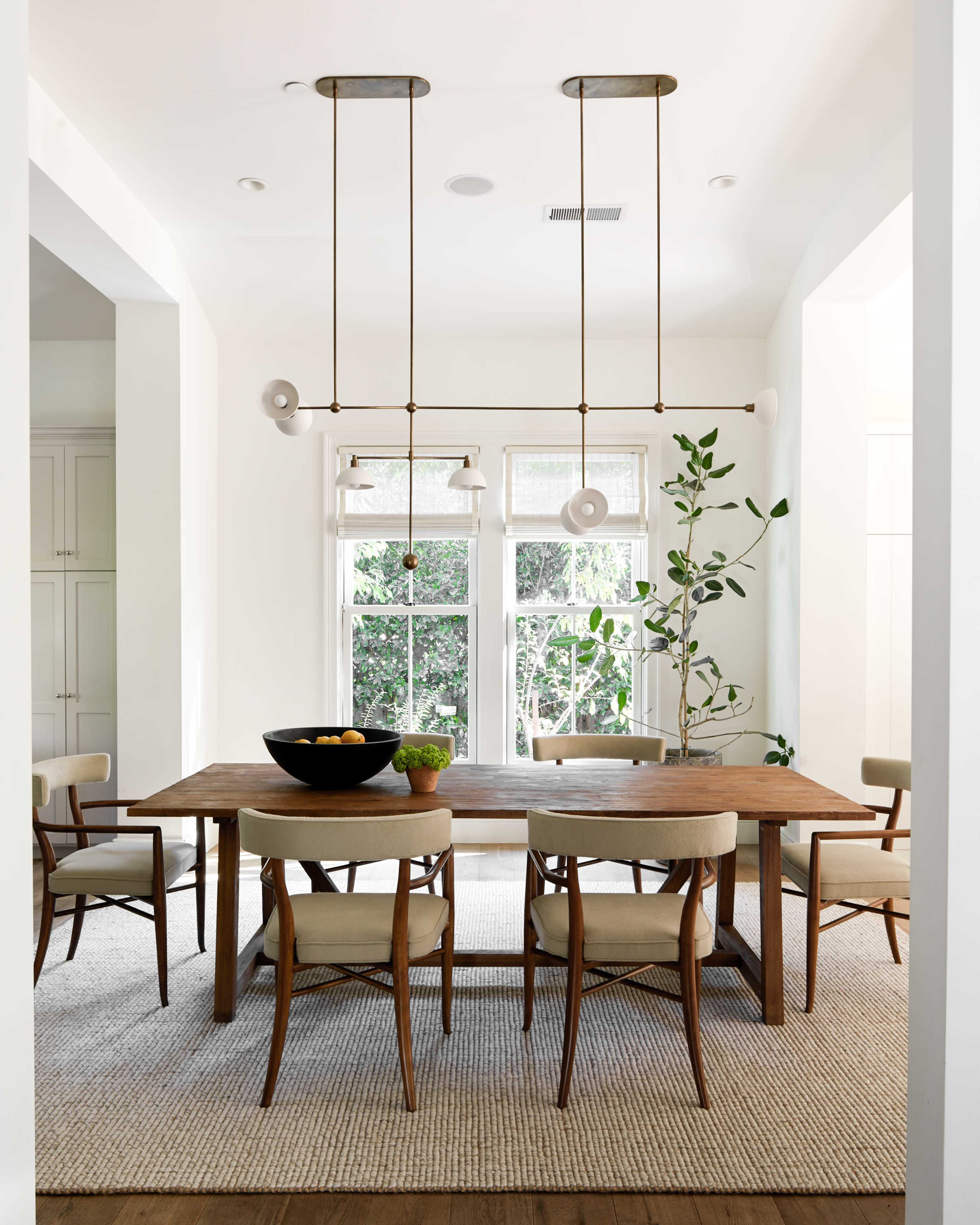 A wooden dining table surrounded by six chairs is situated in a bright, airy room with large windows and hanging light fixtures.