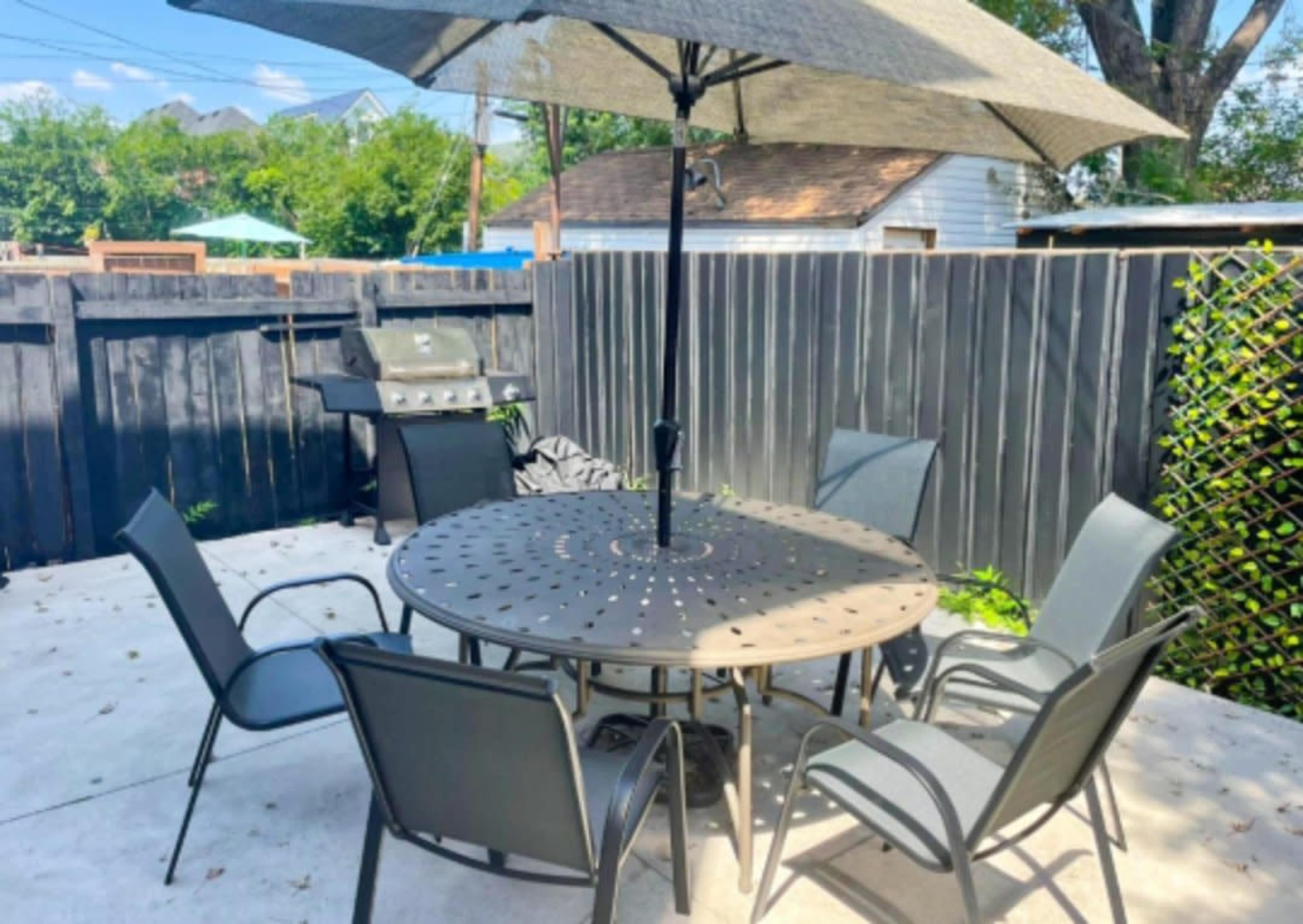 A patio area features a round table with six chairs under a large umbrella, adjacent to a grill and surrounded by a wooden fence.