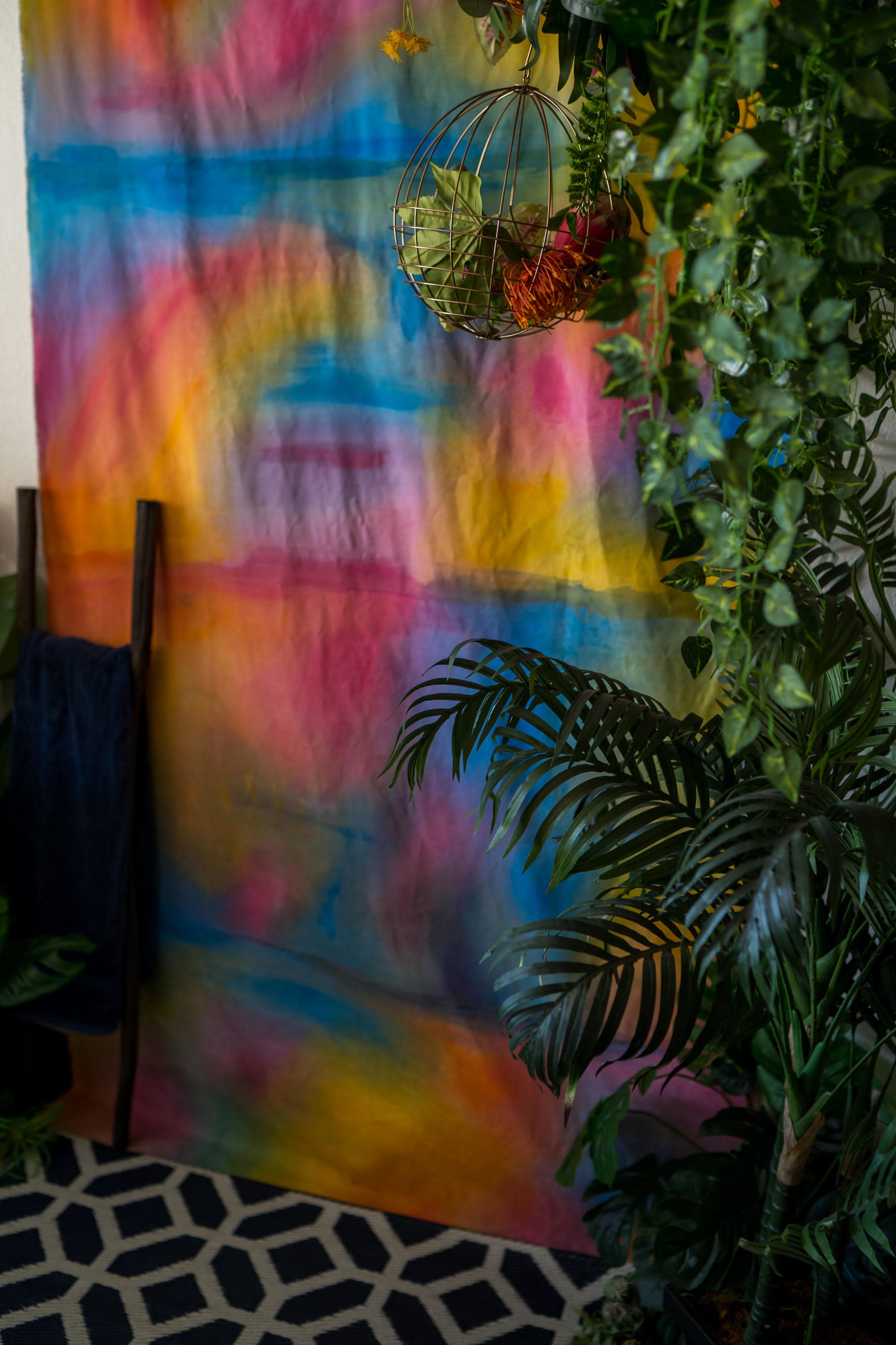 The image features a colorful, abstract backdrop with a hanging planter and lush green foliage in a corner.