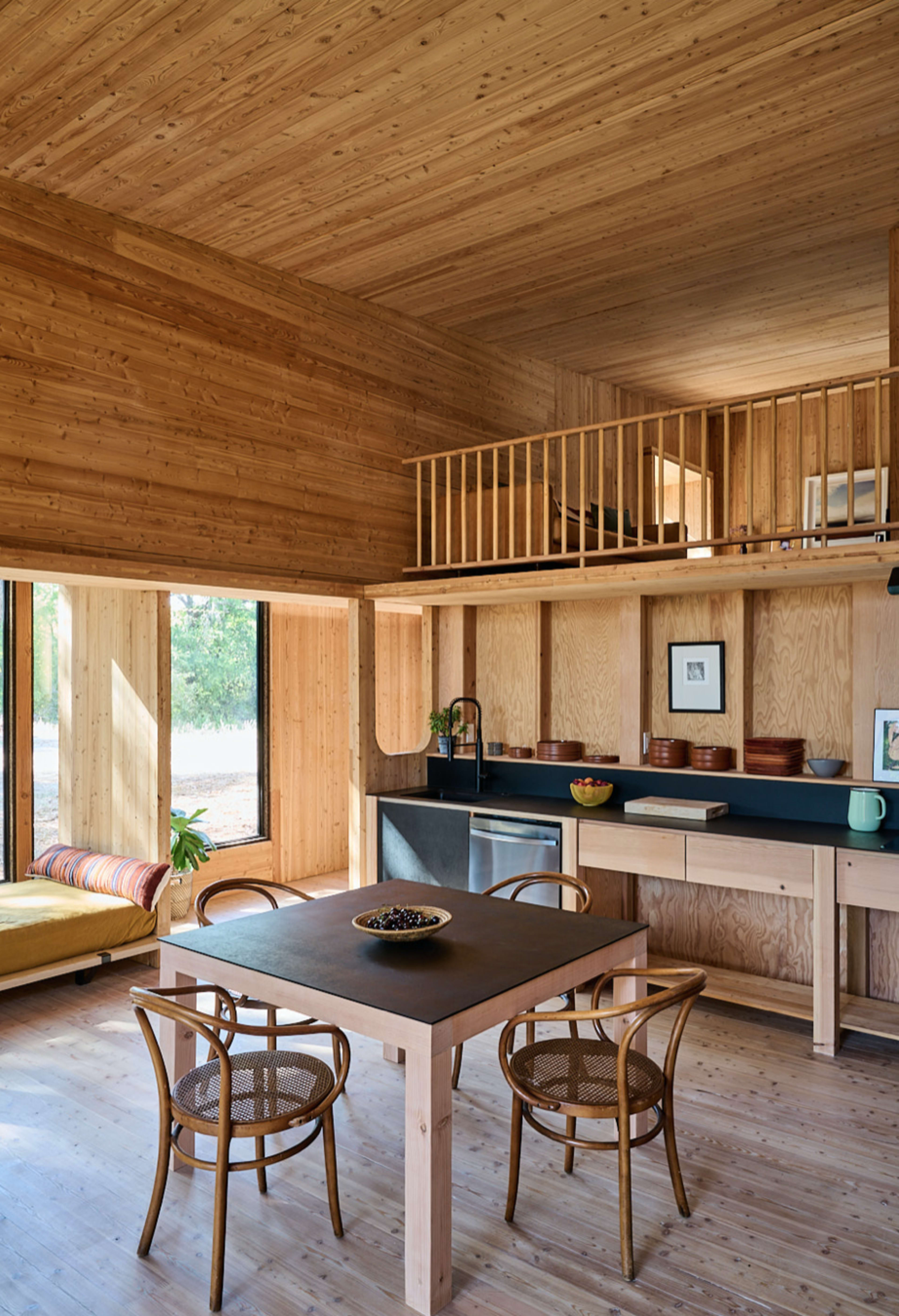 A modern wooden interior featuring a dining table with chairs, a kitchen area with shelves, and large windows that provide natural light.