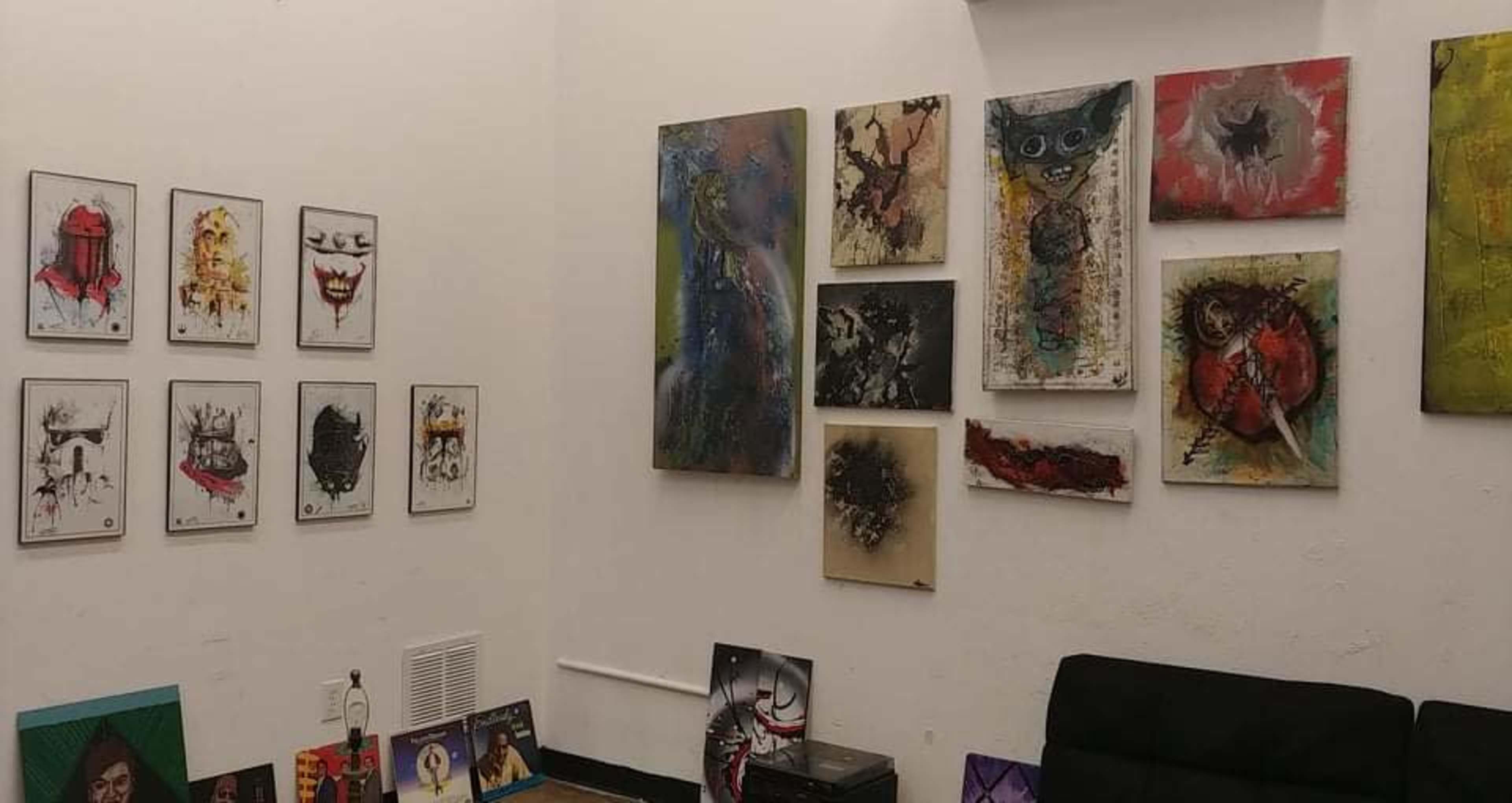 A gallery wall filled with various framed artworks, including abstract and figurative pieces, alongside a dark couch in a simple interior space.