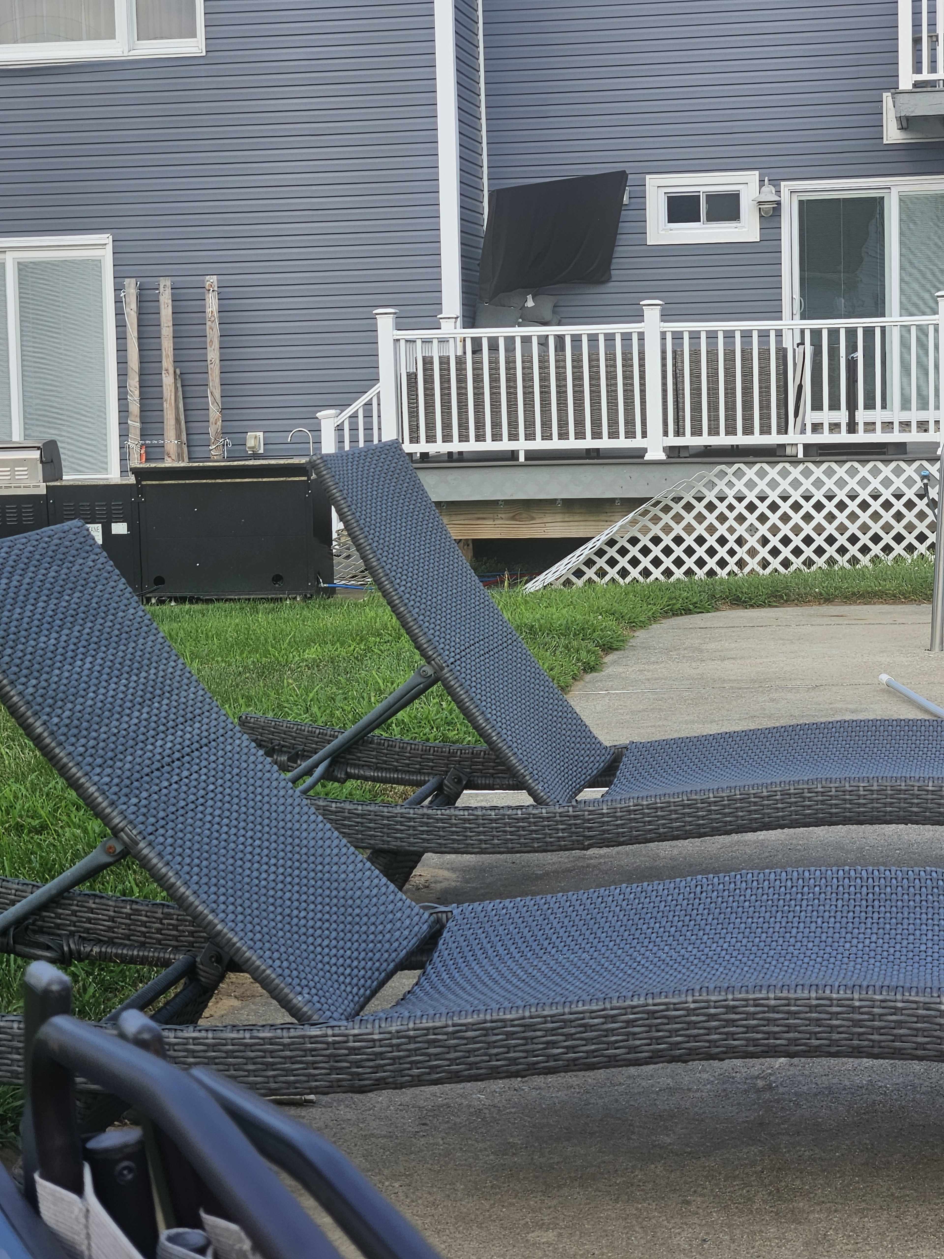 Two chaise lounges are positioned on a concrete patio in front of a house with a blue exterior and a white railing.