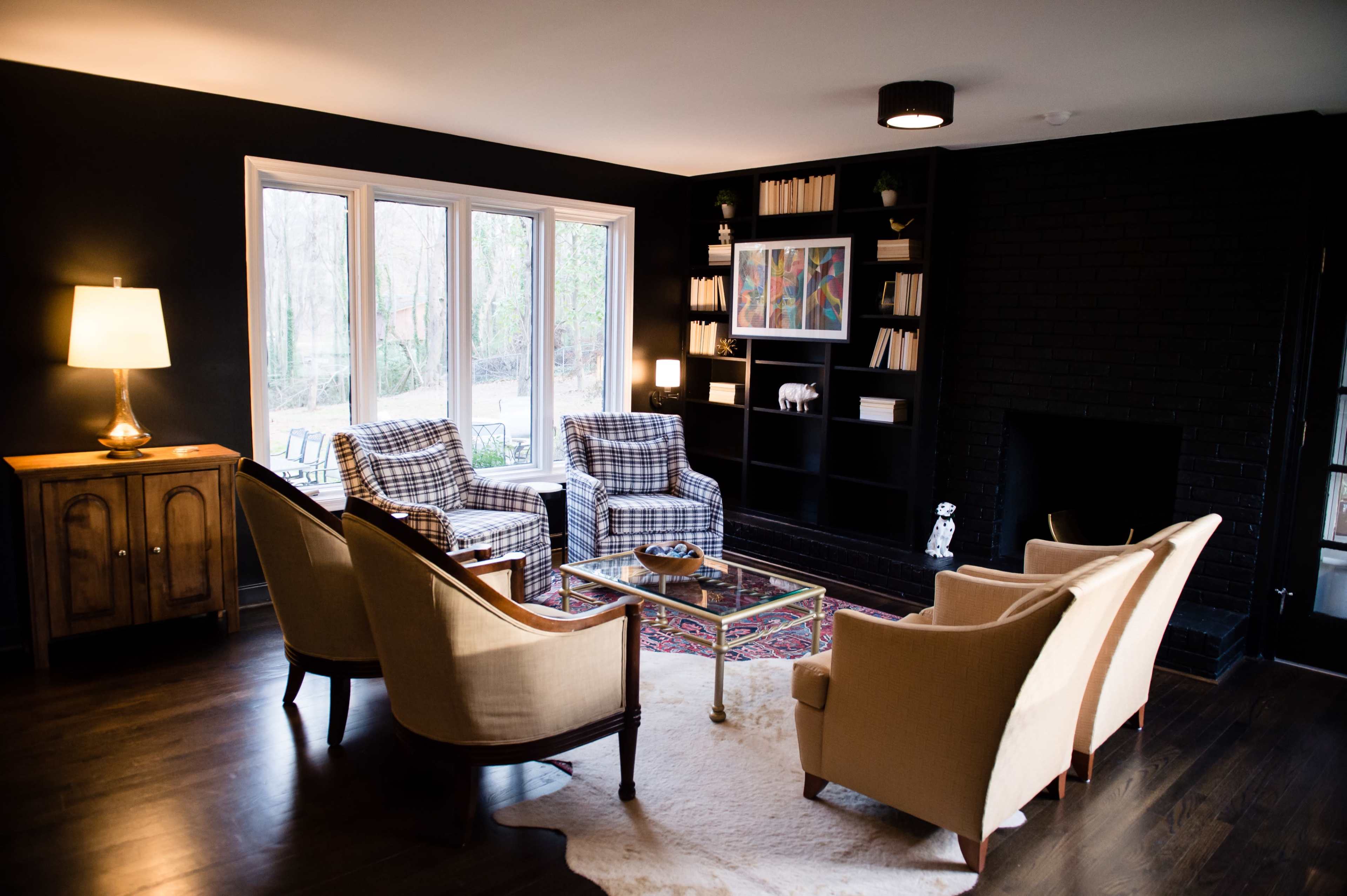 The scene features a cozy living room with two armchairs and two patterned chairs arranged around a coffee table, alongside a bookshelf and large windows overlooking a garden.