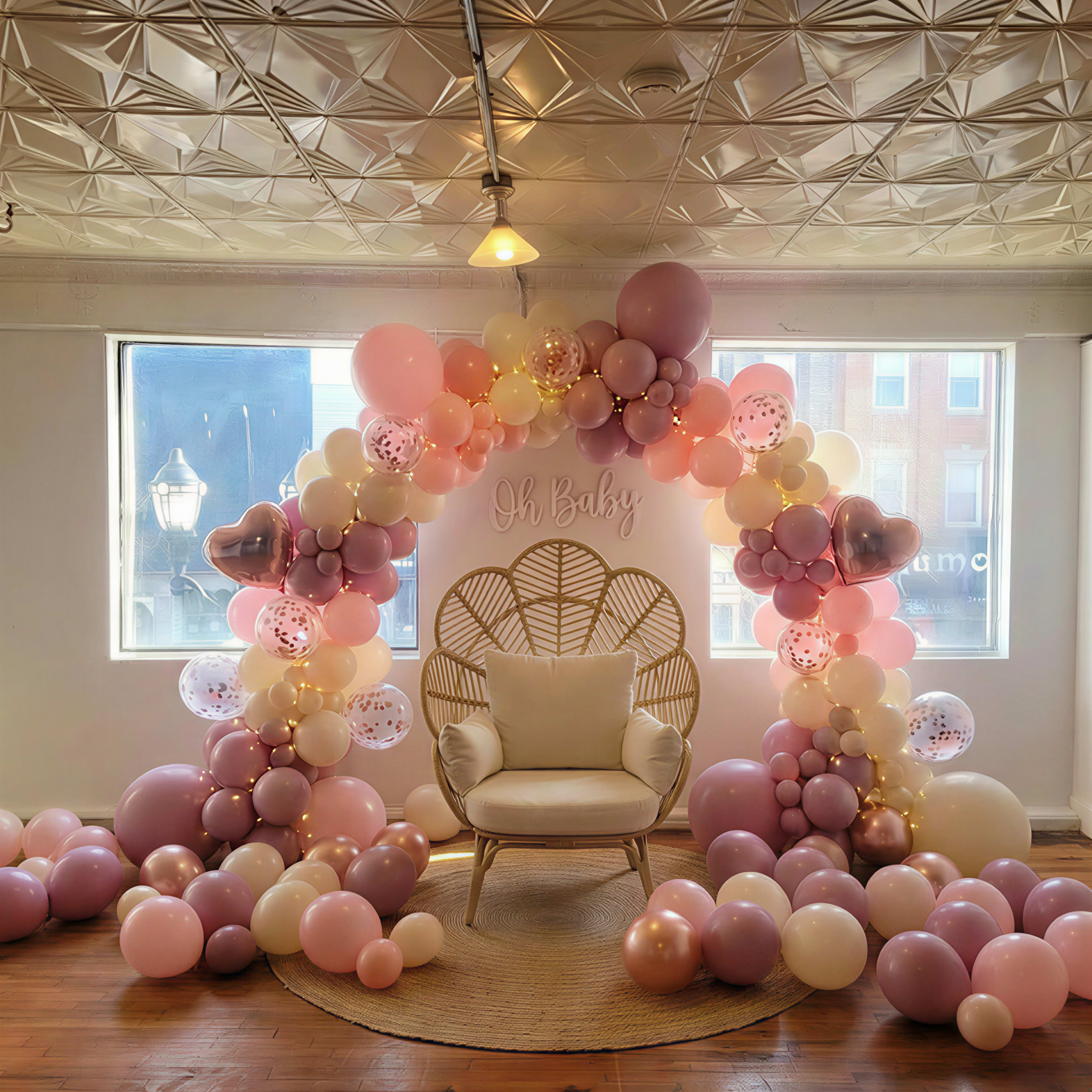 A decorative arch made of pink and cream balloons surrounds a white chair, set against a bright window with the text "Oh Baby" on the wall.