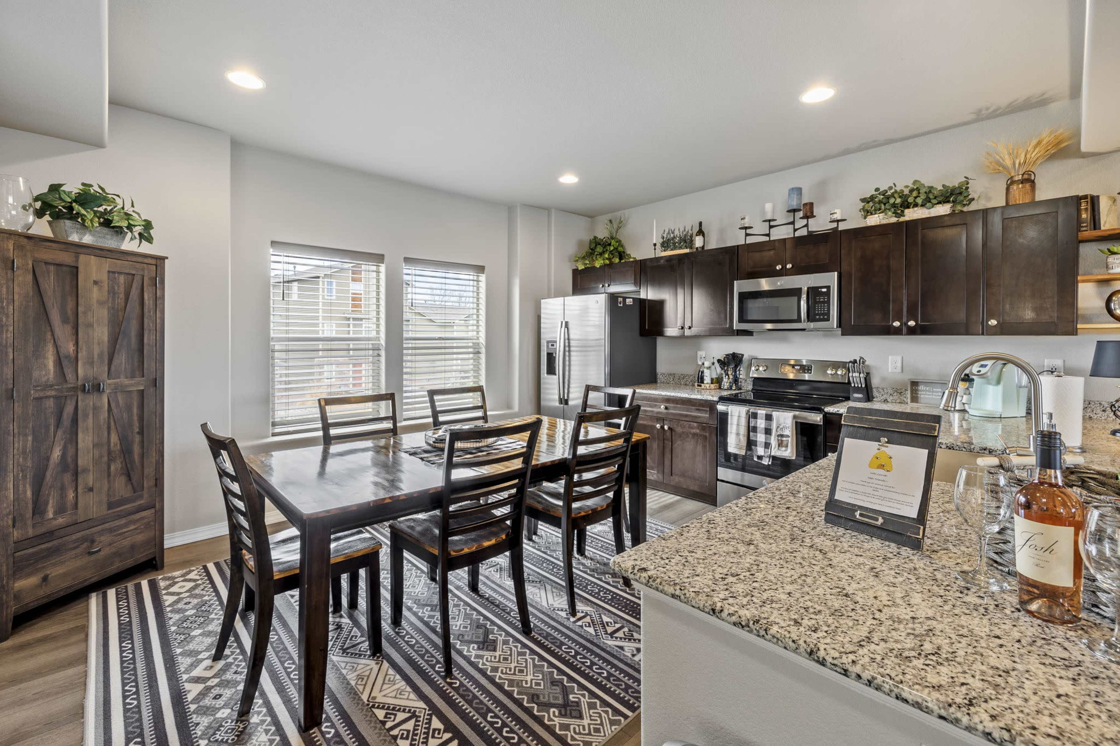 A modern kitchen with dark wood cabinetry, a large dining table, and stainless steel appliances is arranged in a well-lit space with a patterned rug.