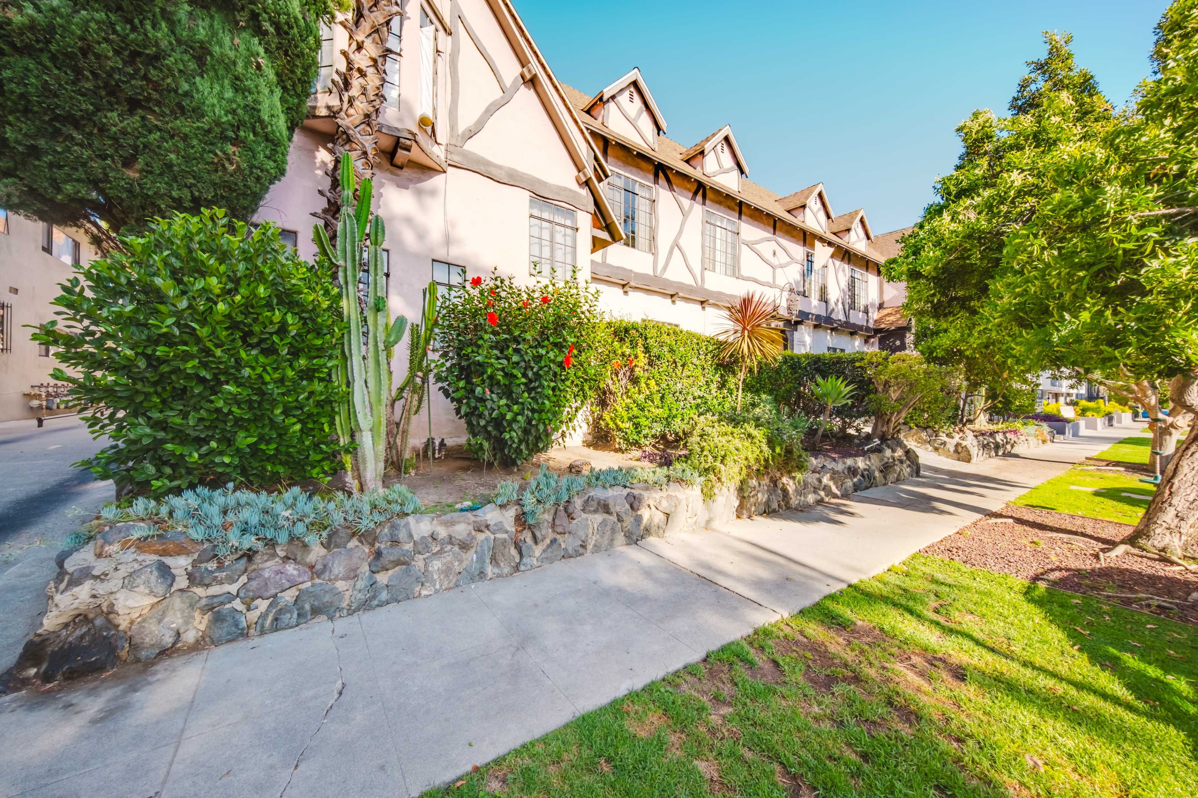 The image shows a walkway lined with green shrubs and plants in front of a brown, multi-story building with a sloped roof.