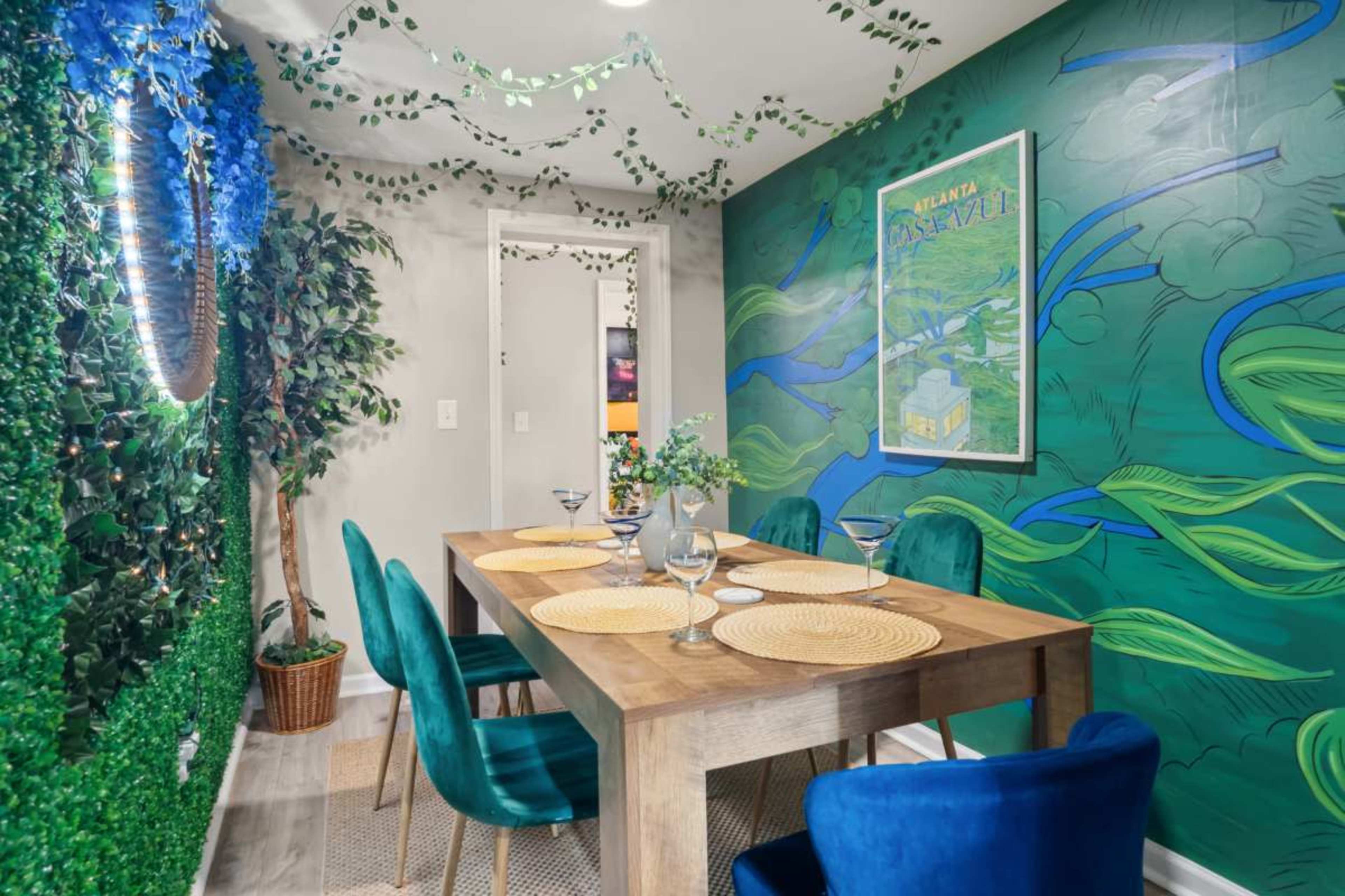 The image shows a dining area with a wooden table set for four, surrounded by green and blue wall art, decorative plants, and soft lighting.