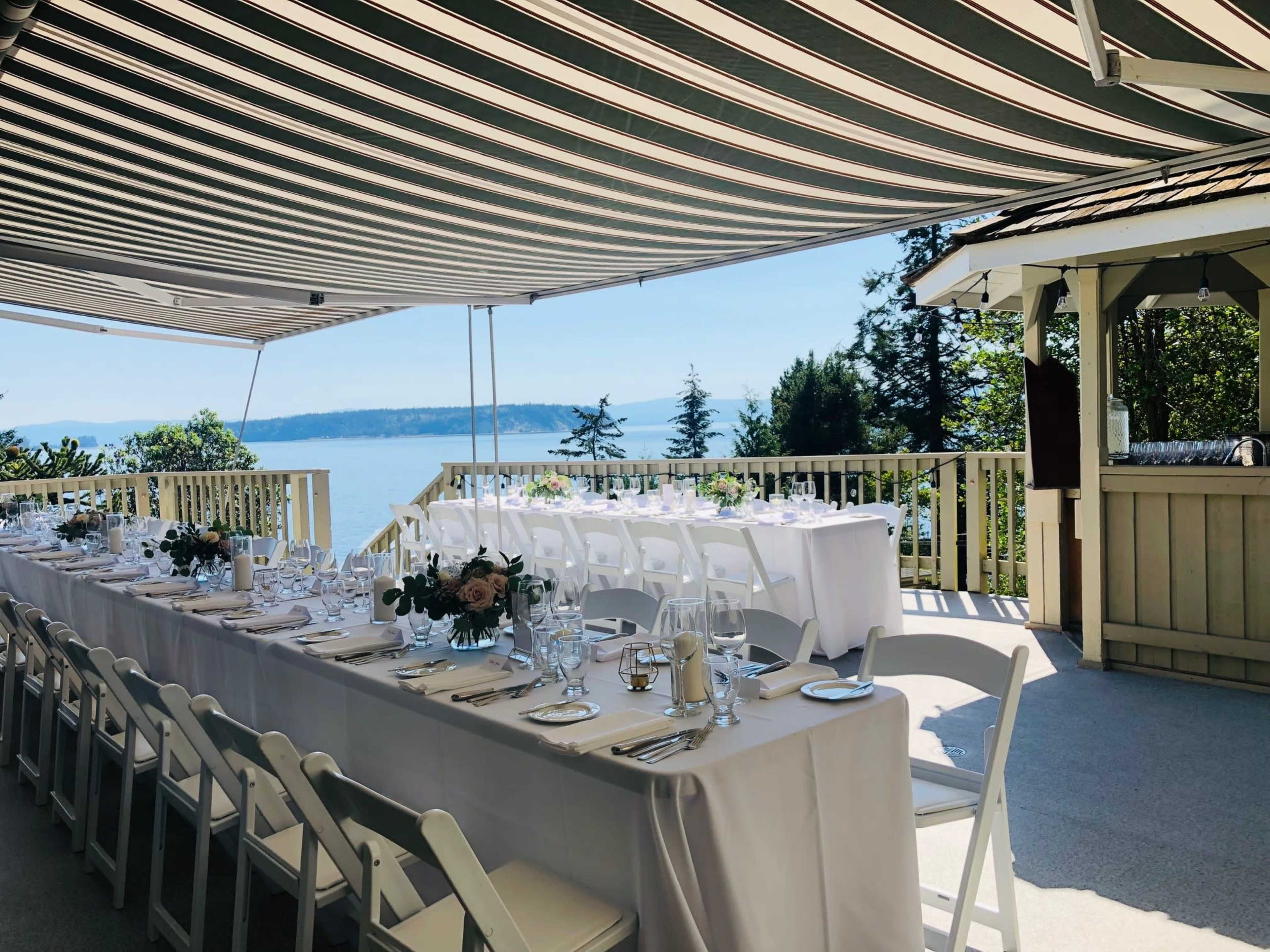 A long dining table is set with tableware and flowers under a striped canopy, overlooking a body of water and trees.