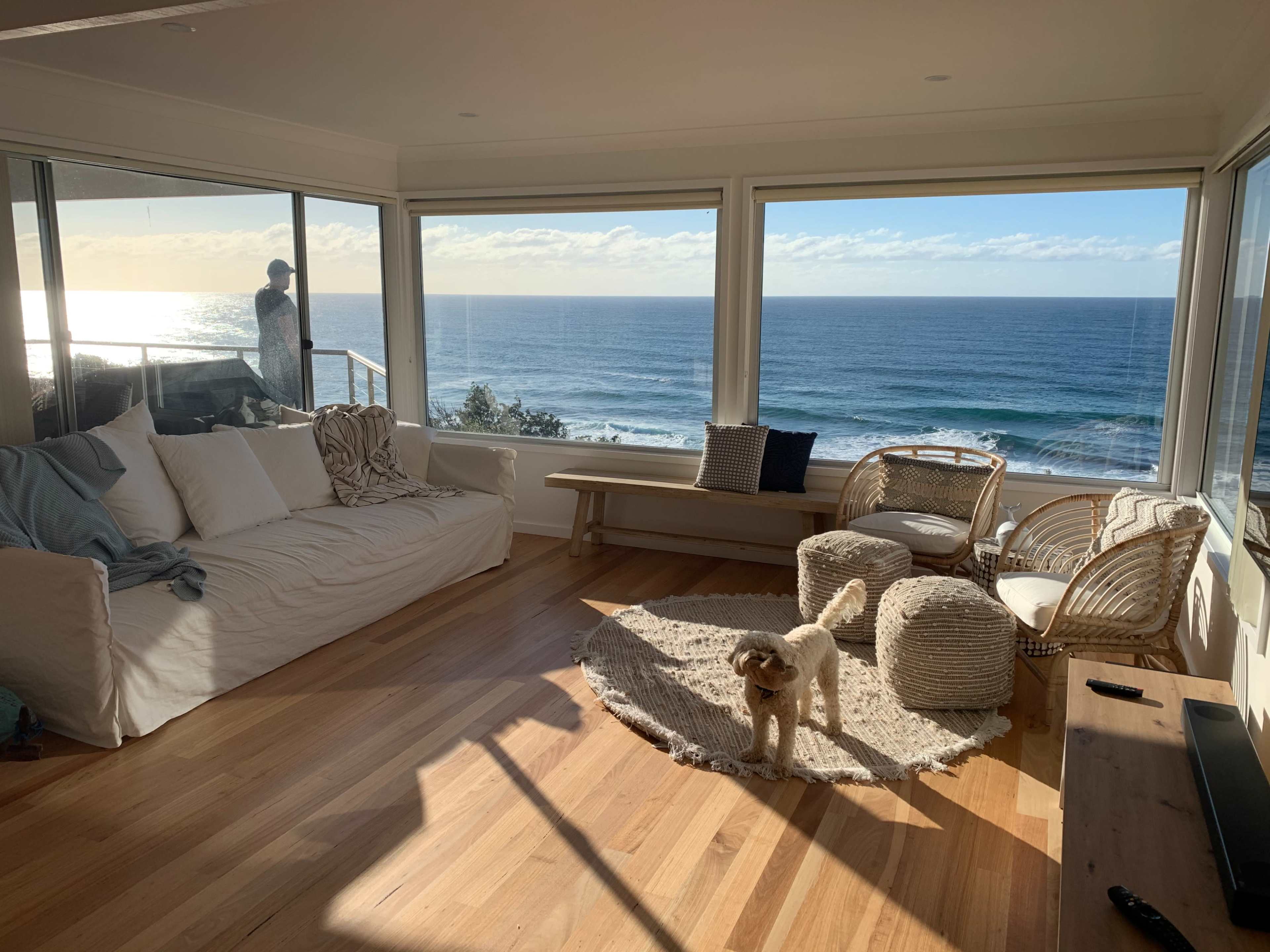A spacious living room with a view of the ocean features a light-colored sofa, woven furniture, and a dog on a circular rug.
