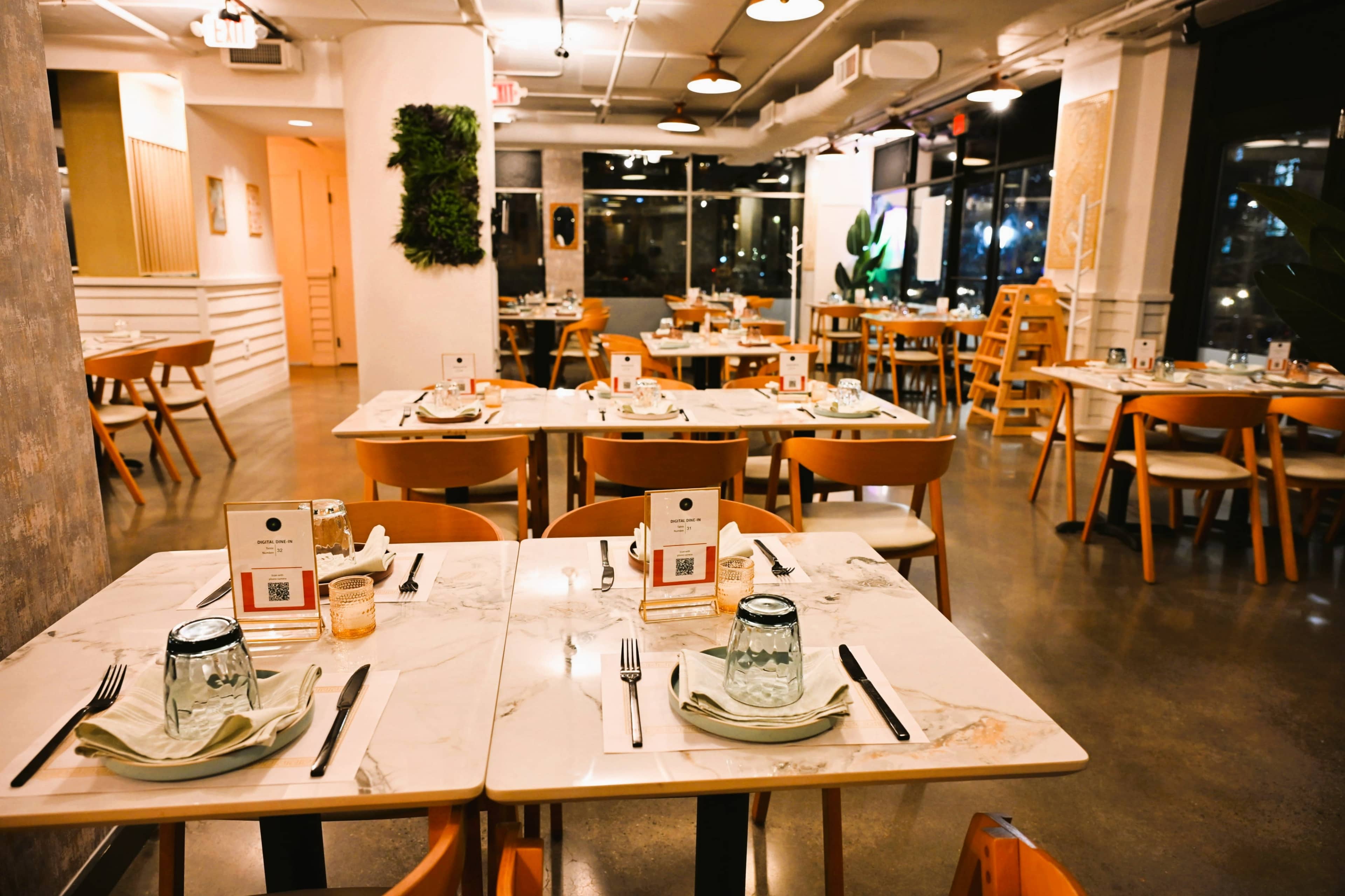 The image shows a modern restaurant interior with multiple tables set for dining, featuring marble tabletops, minimalist decor, and large windows providing natural light.