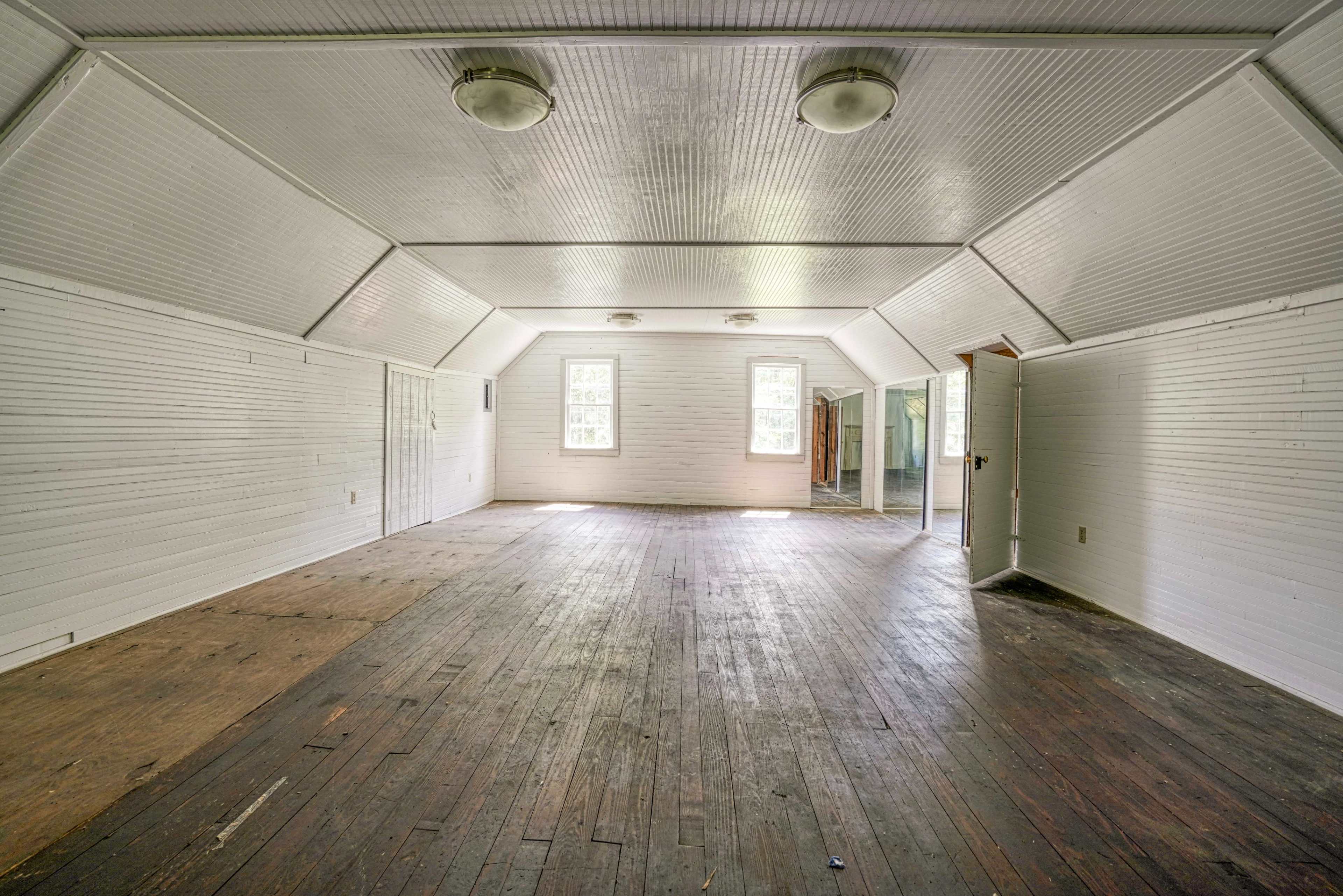 The image shows a spacious, empty room with wooden floors and sloped ceilings, illuminated by overhead lights.