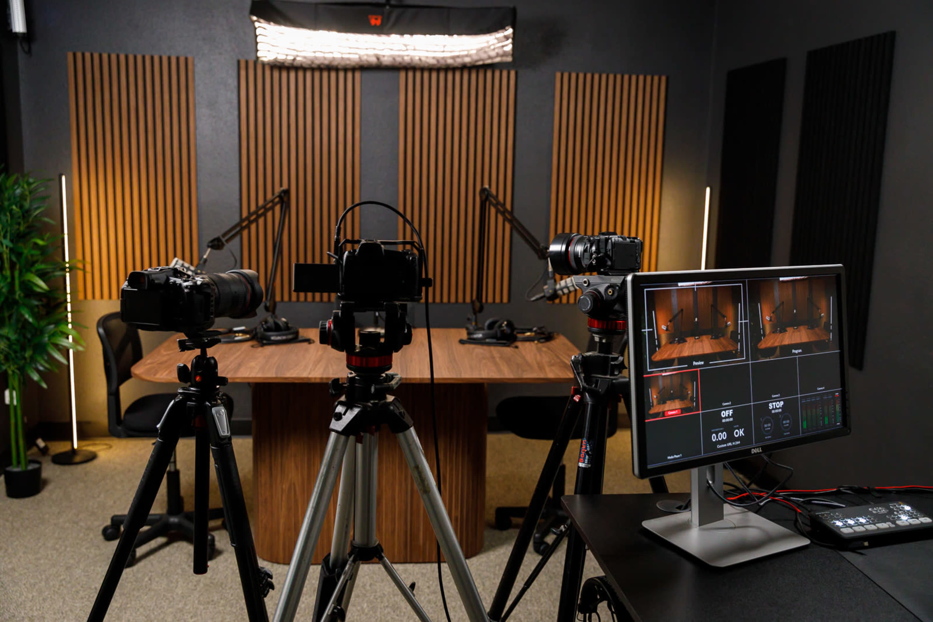 The image shows a recording studio setup with two cameras on tripods facing a large wooden table surrounded by chairs, and a monitor displaying video feeds.