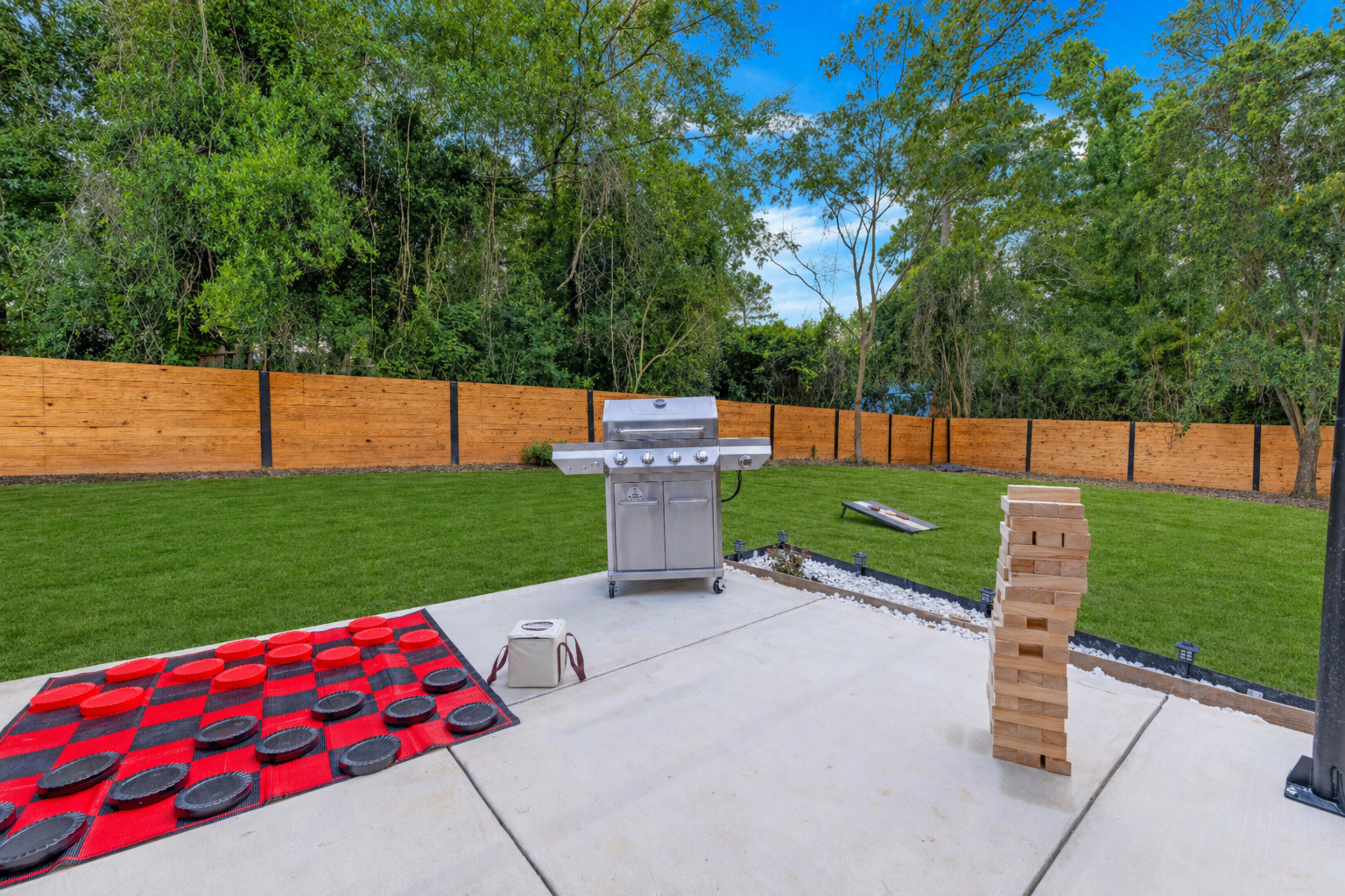 The scene features a backyard patio with a grill, a checkers game setup on a red and black mat, and a stacked game of giant Jenga, all surrounded by a wooden fence and greenery.