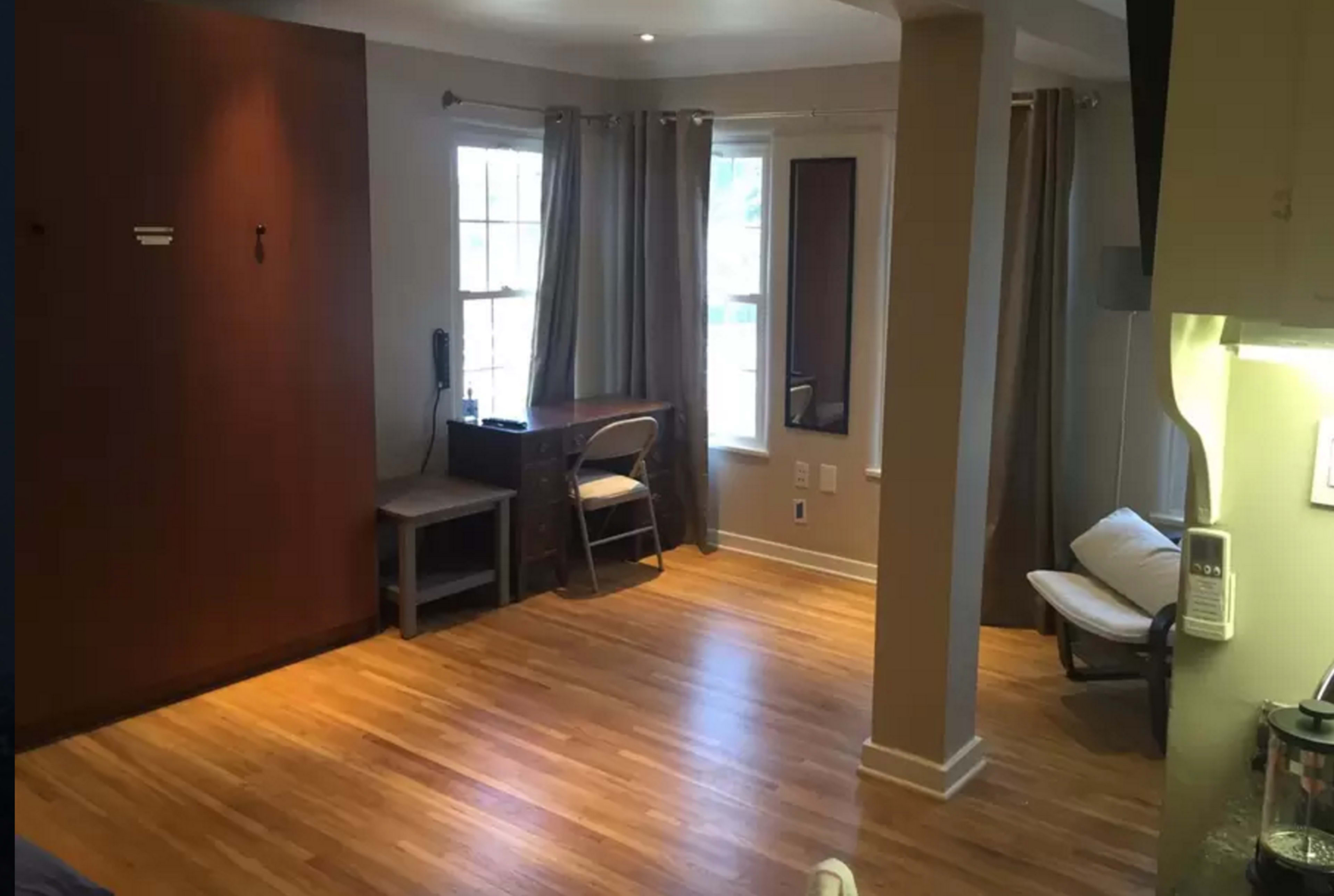 A room with hardwood flooring, a desk and chair near a window, and a wall-mounted storage unit.